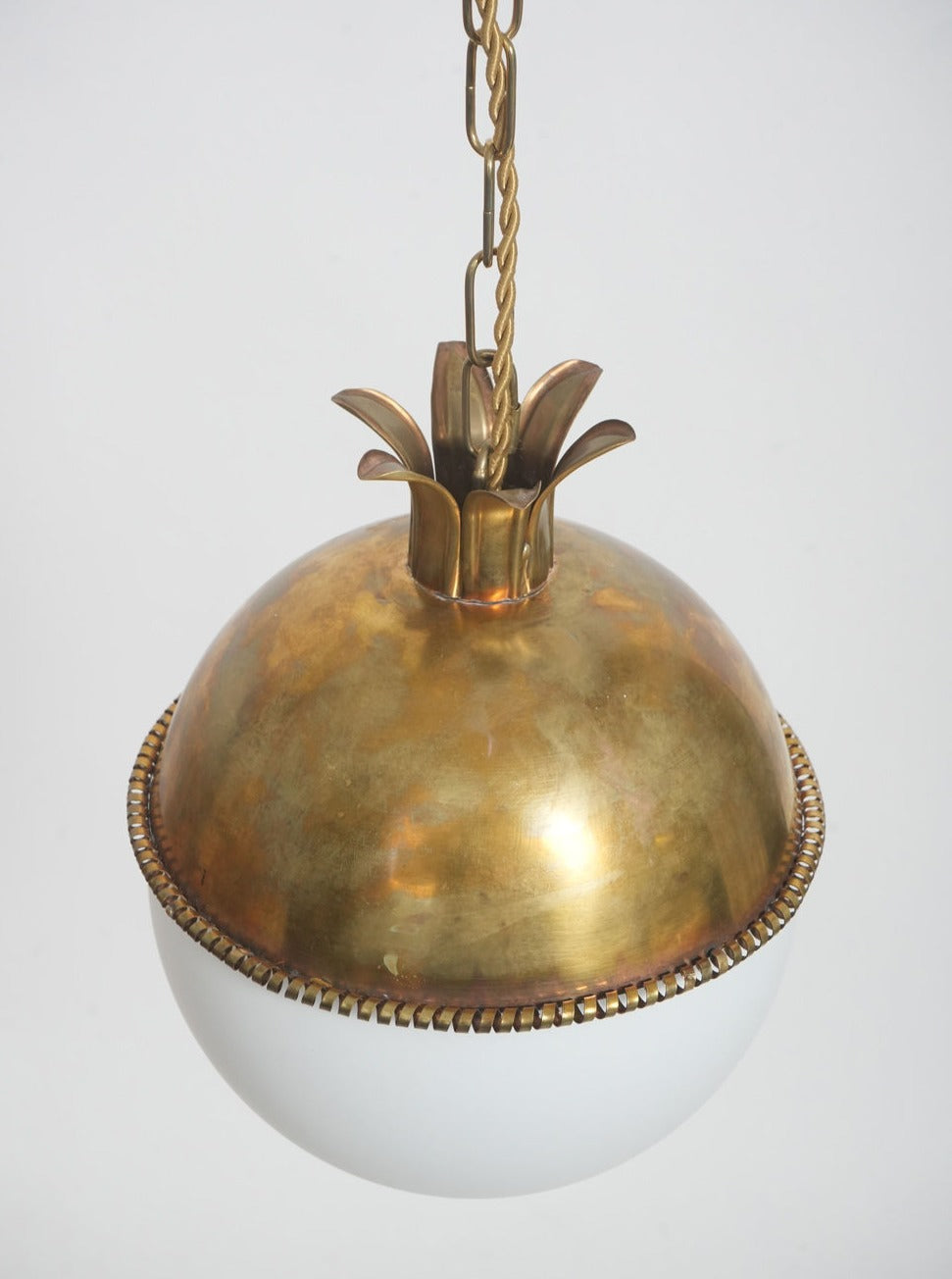 The Granada Ceiling Pendant Lamp by Barracuda Interiors features a brass fixture inspired by a pineapple crown, supporting a round white glass globe with intricate decorative edging.