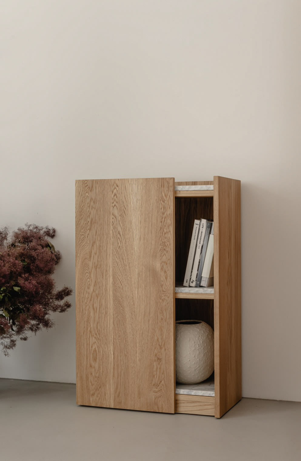 The PAVO Cabinet by un'common, crafted from light solid wood, stands against a beige wall with its door ajar to display books and a decorative vase. A bundle of dried flowers rests on the floor to the cabinet's left.