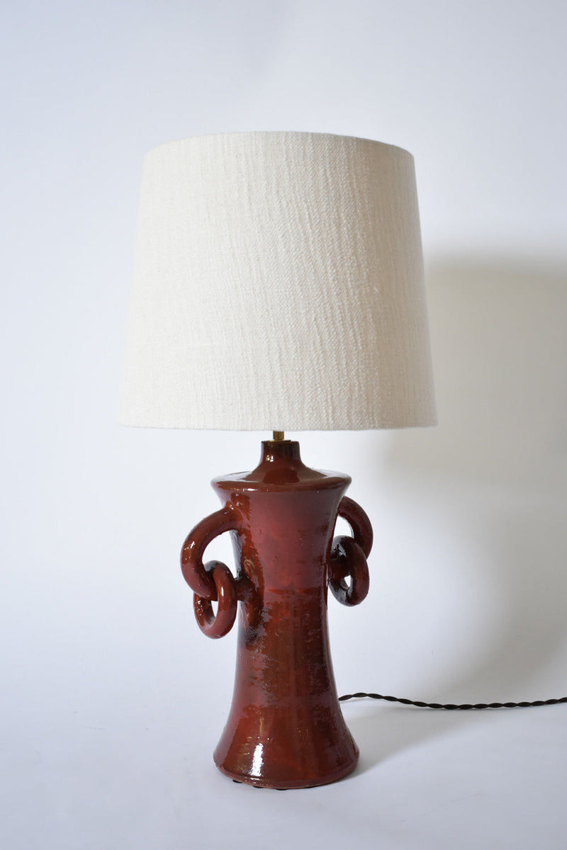 The "Kasaï" Red Handles Ceramic Table Lamp by Barracuda Interiors features a glossy reddish-brown ceramic base with sculpted circular handles, a large off-white fabric shade, and a twisted black cord. Made in Portugal.