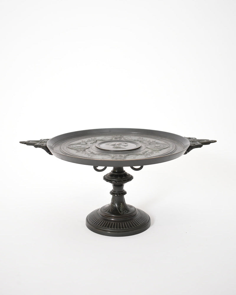 The Barbedienne Style Pedestal Cup, XIXth c. by Barracuda Interiors features ornate dark metal, twin handles, and engraved patterns on a round shallow dish with antique patina, displayed against a plain white background.