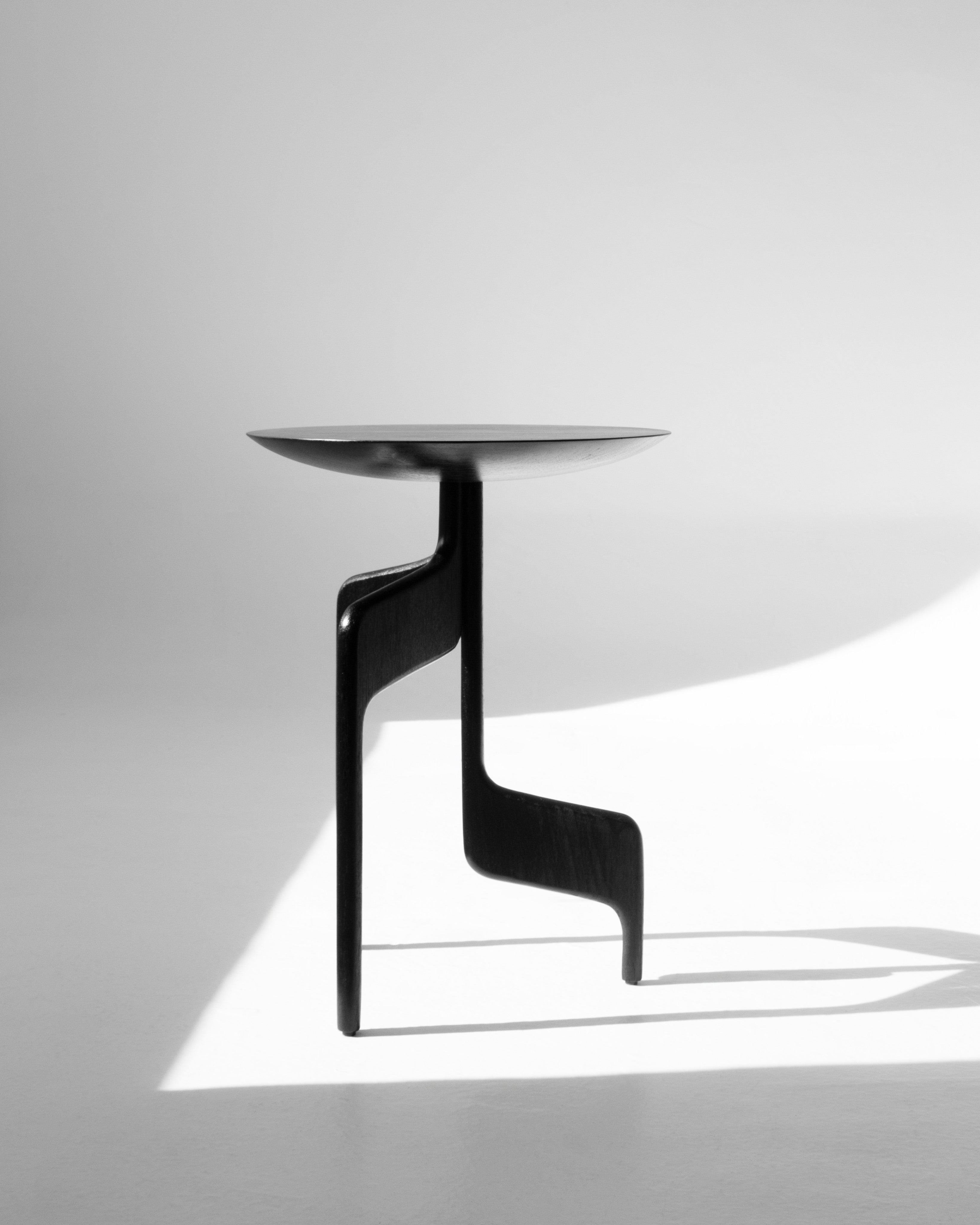 The "Vereda" Side Table by Luciano Costa Estúdio features a modern, minimalist black silhouette with an abstract sculptural form, casting a striking shadow on a white background under bright, directional lighting.
