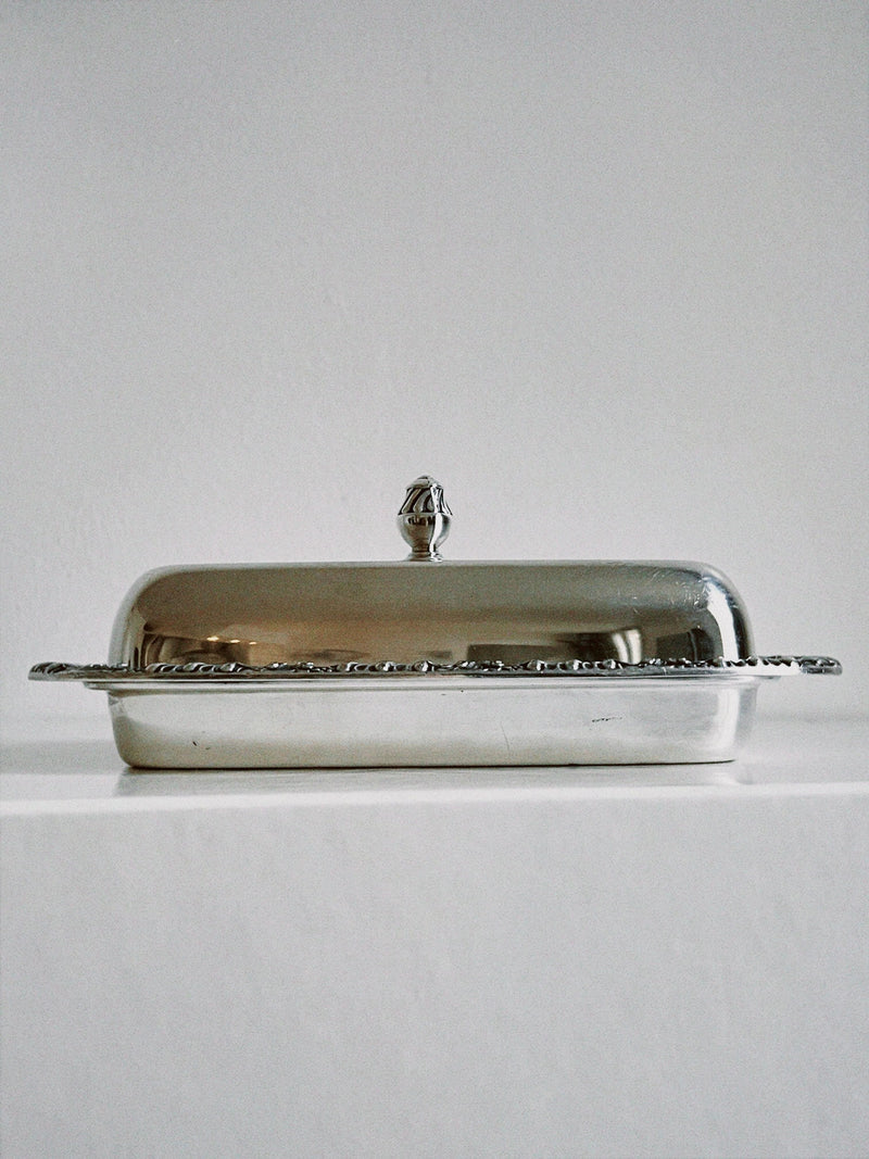 The Manushi Silver Ornate Butter Dish, featuring a decorative handle on its lid, is displayed on a white surface against a plain white background.