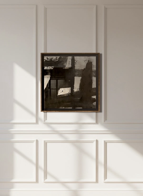 “Marblehead Harbor,” an ink and charcoal on raw canvas by Médecine, hangs in a dark frame on a white paneled wall, with sunlight casting soft, diagonal shadows across the wall and floor.