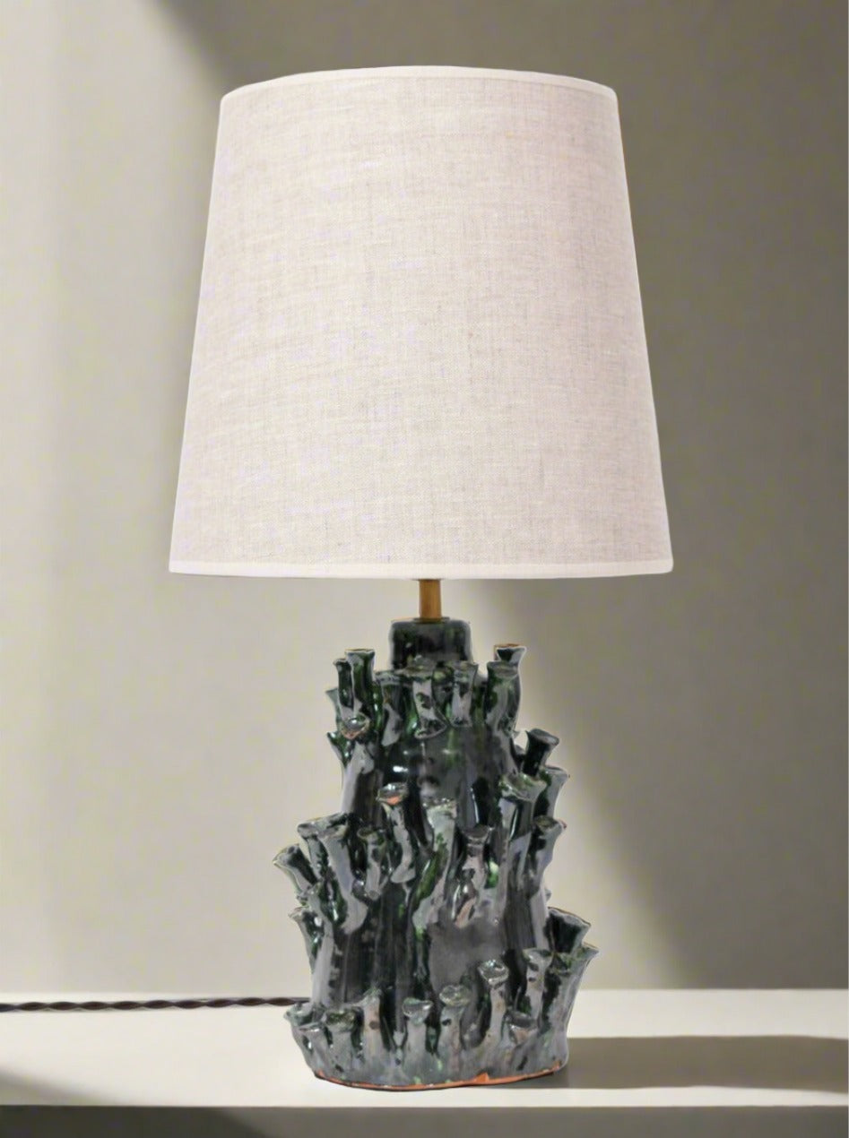 The "Belize" Green and Black Lamp by Barracuda Interiors, featuring a textured sculptural ceramic base and a light fabric shade, made in Portugal, sits on a white surface against a neutral background.