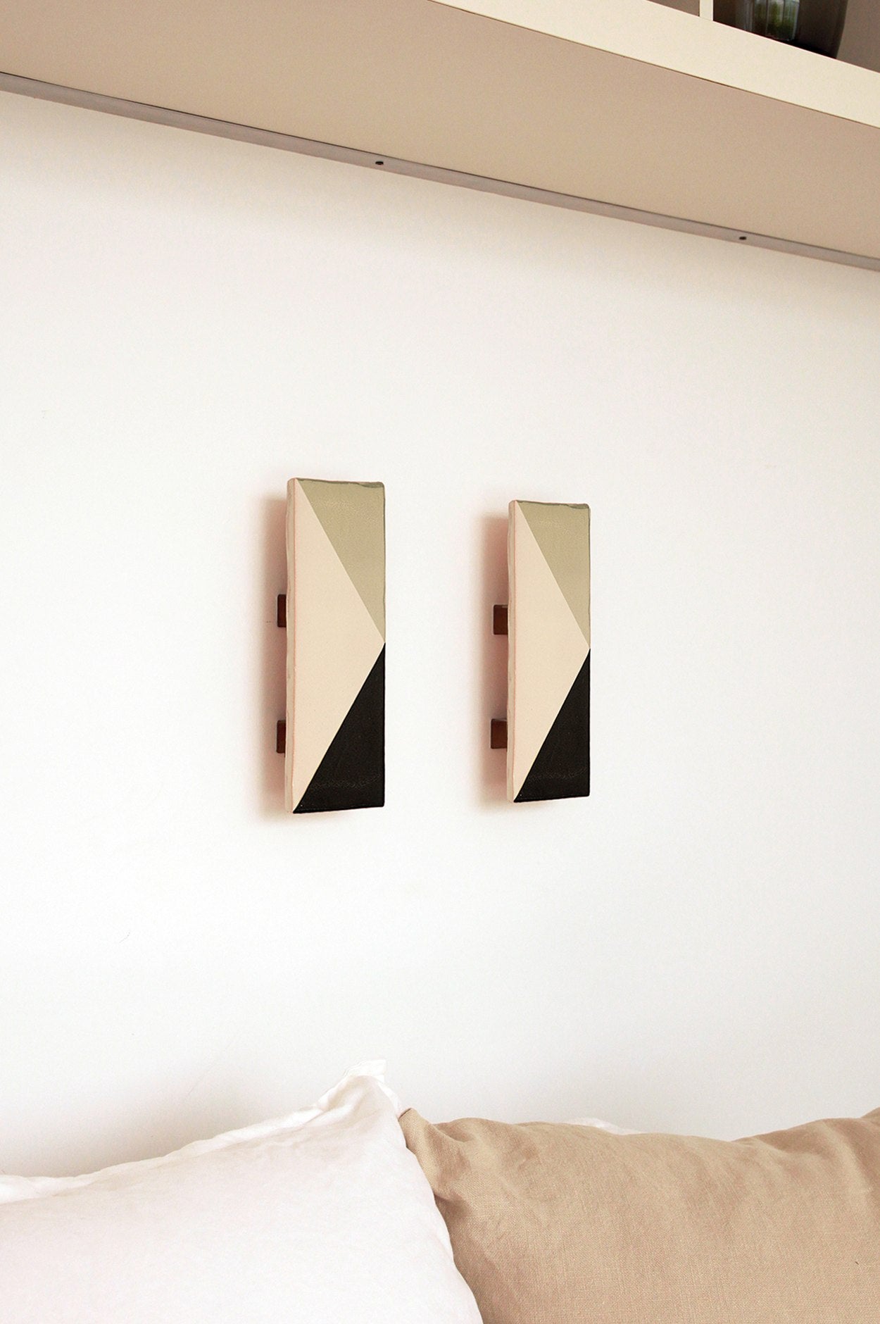 Two Tile VN appliques murales by Violaine d'Harcourt, featuring angular beige, cream, and black ceramic patterns, hang side by side on a white wall above a beige and white sofa. A light-colored shelf is visible at the top of the image.