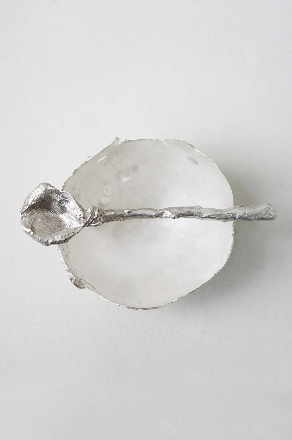 The L34 - Small Bowl in Silver by Completedworks, featuring an irregular edge, is paired with a matching textured silver spoon and photographed from above on a light gray surface.