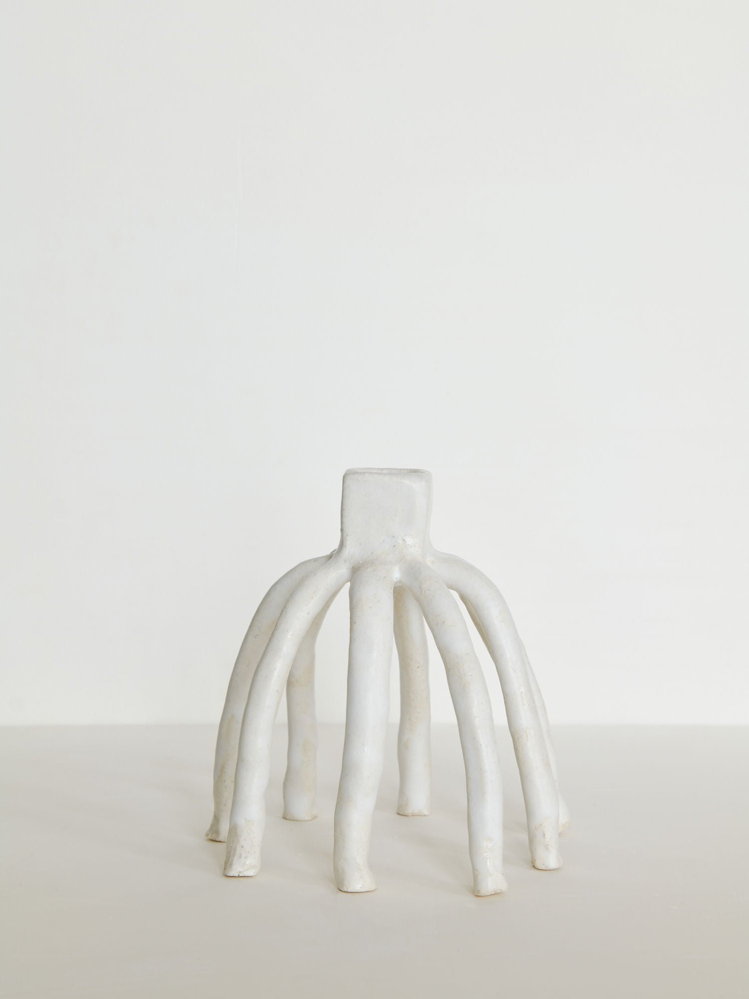 The Candleholder La Chose 1 by Claire Pain is a white ceramic sculpture featuring six elongated, curved legs radiating from a central cylindrical base, resembling an abstract candle holder against an off-white background.