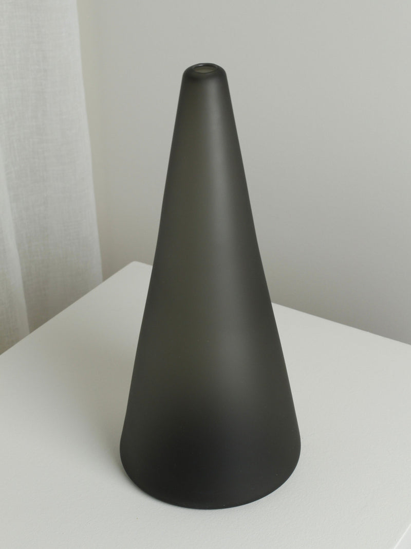 The XLarge Vase in Smoke by Verre d'Onge, featuring a matte black cone shape and a smooth finish with a small circular top detail, sits on a white surface near a white wall and sheer curtain.