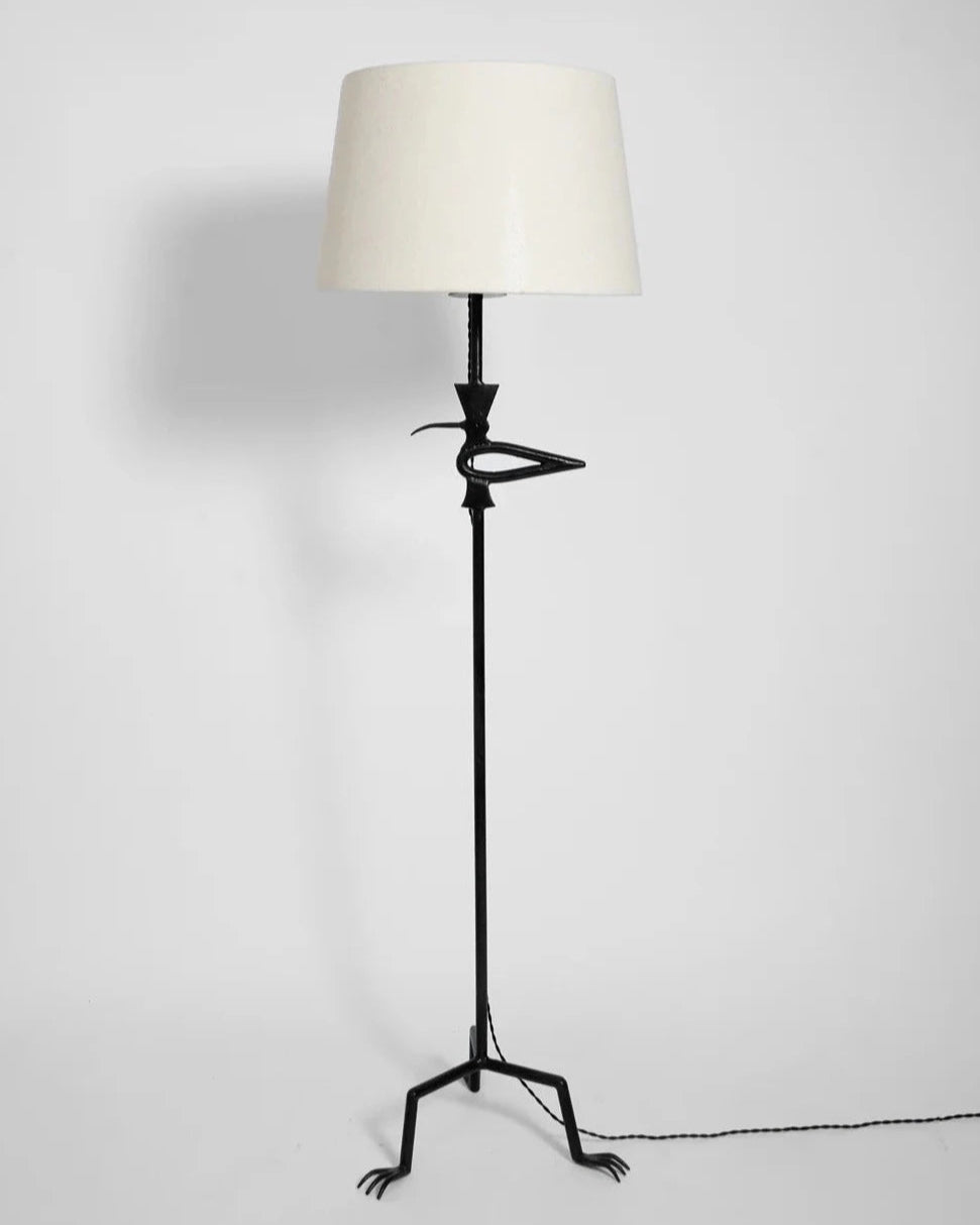 The "Brejos" Floor Lamp by Barracuda Interiors features a white shade and black wrought iron stand shaped like a bird, complete with bird-like feet and a beak, and offers customizable lighting against a plain white background.