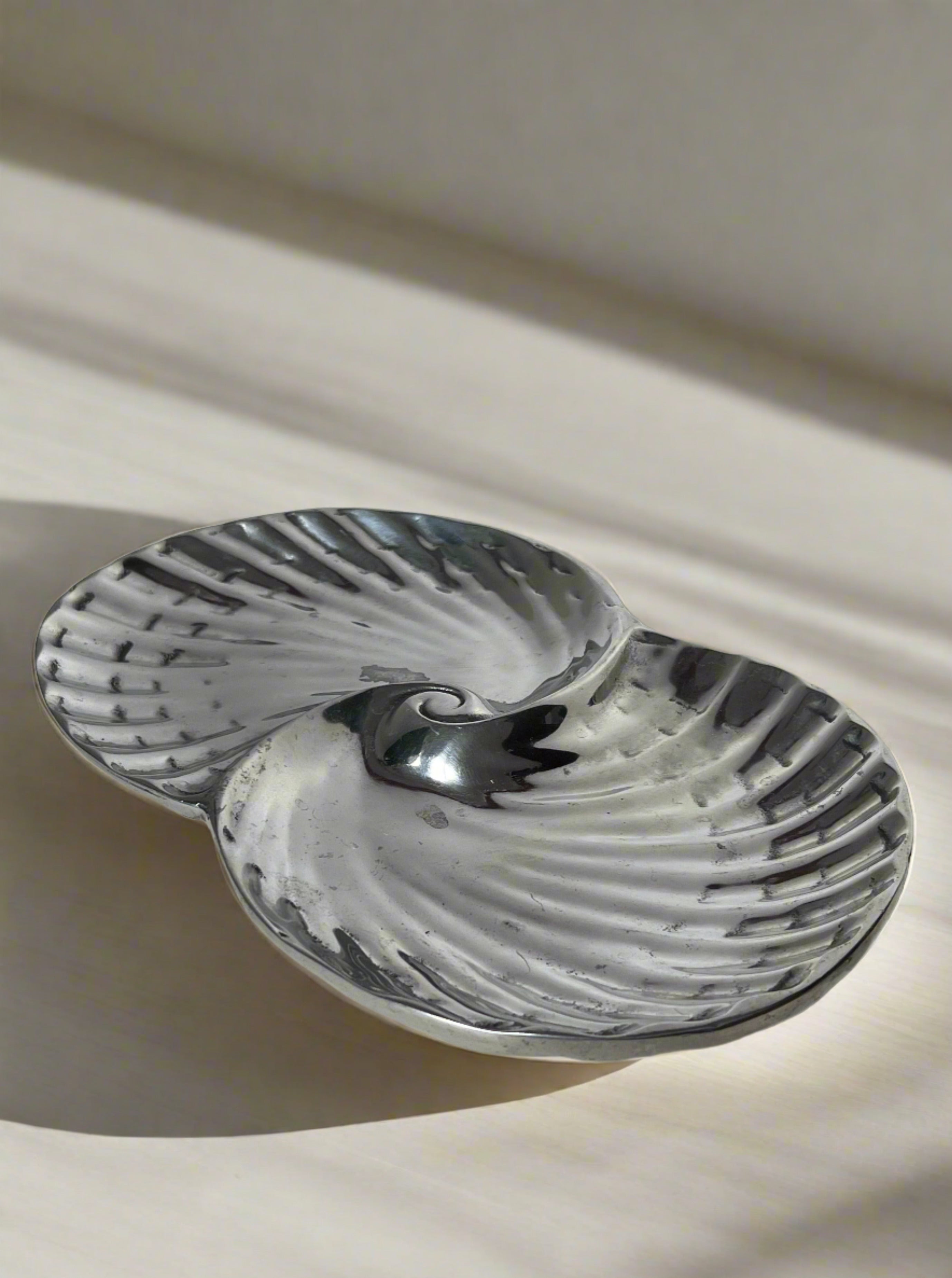 The Manushi Swirl Decorative Dish features a silver, shell-shaped design with two connected sections and rests on a light surface, casting gentle shadows in natural light.