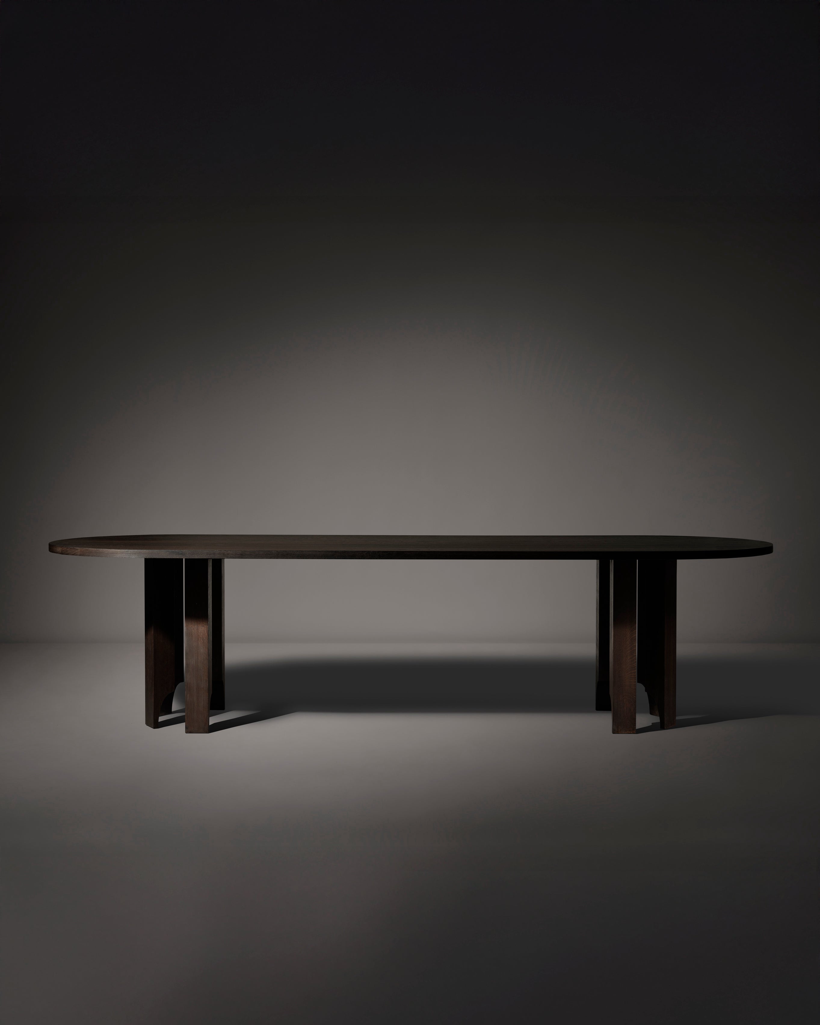 The Mokko Acer Capsule Table, made from solid European oak with rounded edges and sturdy rectangular legs, stands alone in a dimly lit, empty room with a plain background.