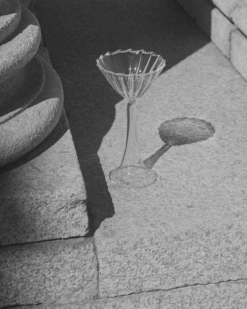 The Campanula — Clear Cocktail Glass by Agustina Bottoni, handmade in Italy, rests empty on a sunlit stone step, casting a striking shadow amidst textured stone and soft natural light.