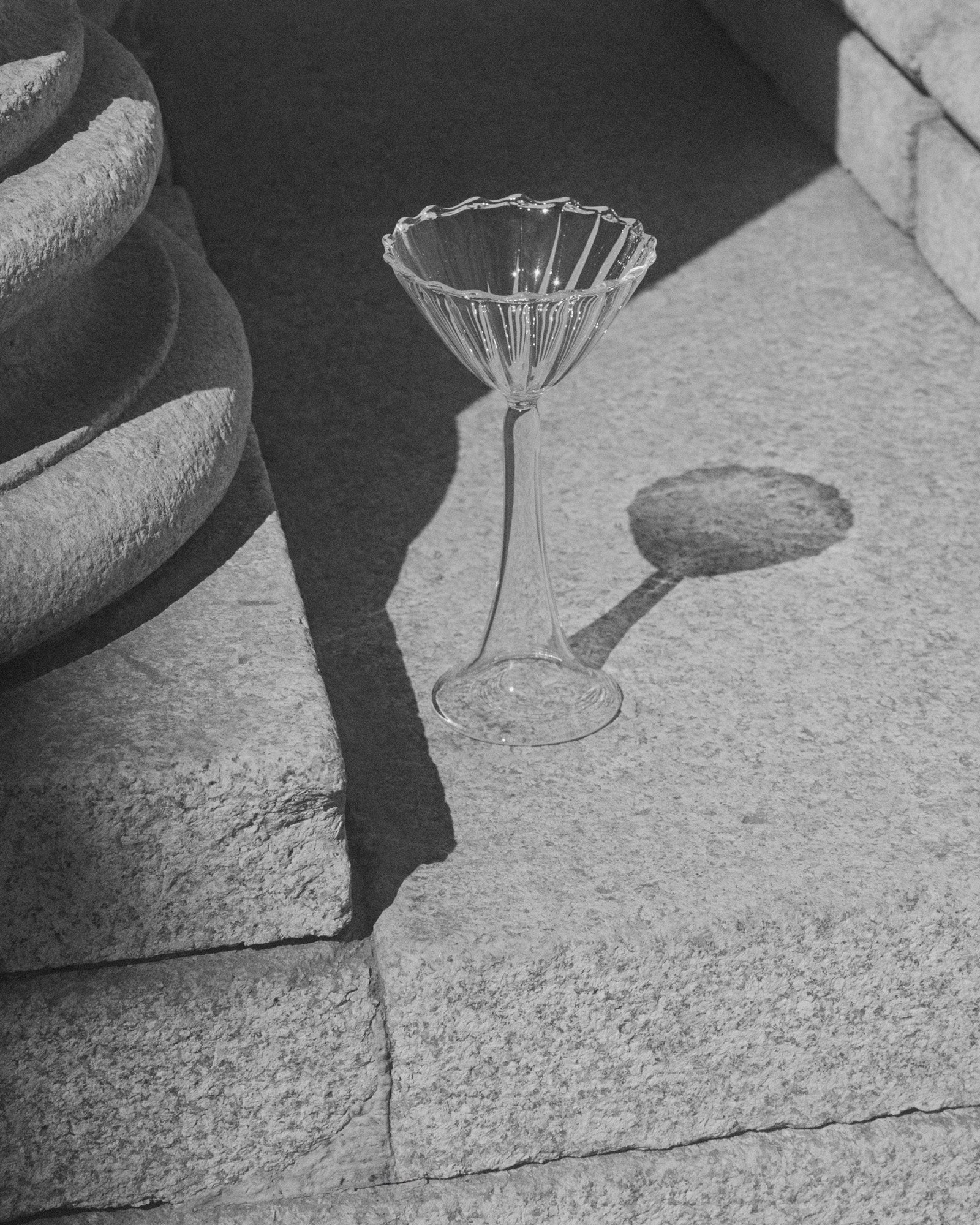 The Campanula — Clear Cocktail Glass by Agustina Bottoni, handmade in Italy, rests empty on a sunlit stone step, casting a striking shadow amidst textured stone and soft natural light.