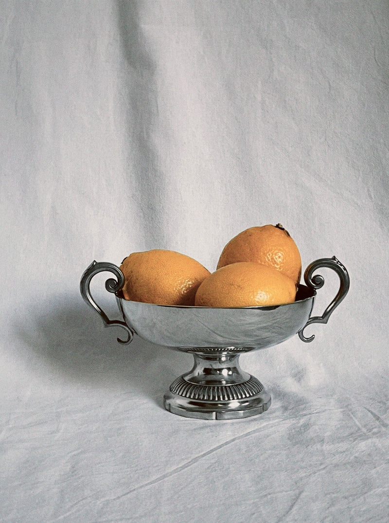 The Manushi Silver Fruit Bowl with ornate handles holds three grapefruits, displayed on a light grey fabric background.
