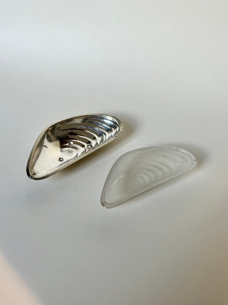 A Manushi Mussel Shell Butter Dish in shiny silver is displayed next to a clear glass mussel shell, both set against a plain white background.