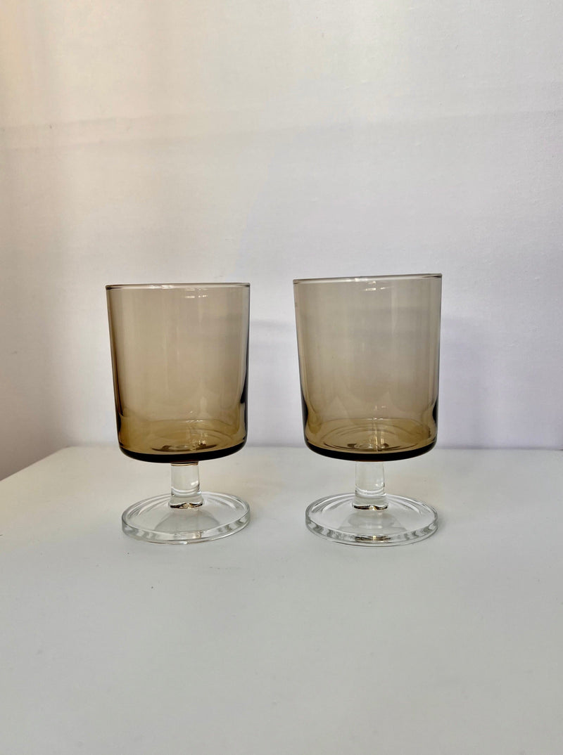 The Manushi Set of Tinted Stem Glasses features two short-stemmed glasses with brown bowls and clear bases, displayed side by side on a white surface with a plain light background.