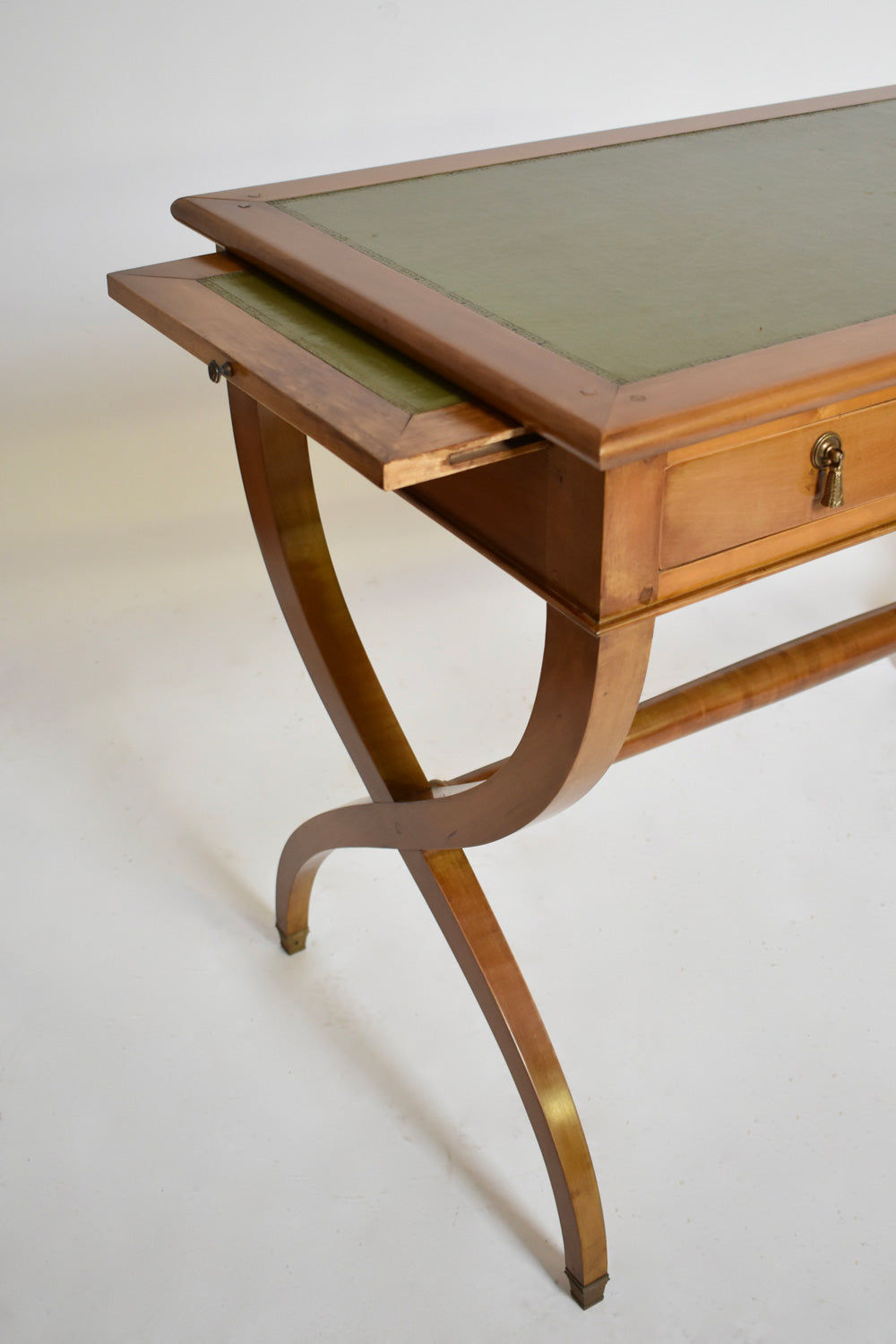 The Directoire style curule console table by Barracuda Interiors (1970s) features curved legs, a green leather writing surface, and an extended sliding panel beneath, displayed against a plain background.