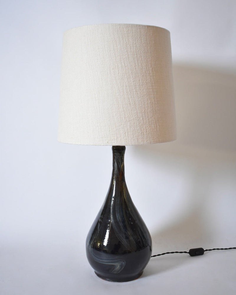The Barracuda Interiors Black Ivory Brush Lamp, Helder, features a dark ceramic rounded base and a cream fabric shade. Set on a plain white background, its visible power cord rests on the surface.