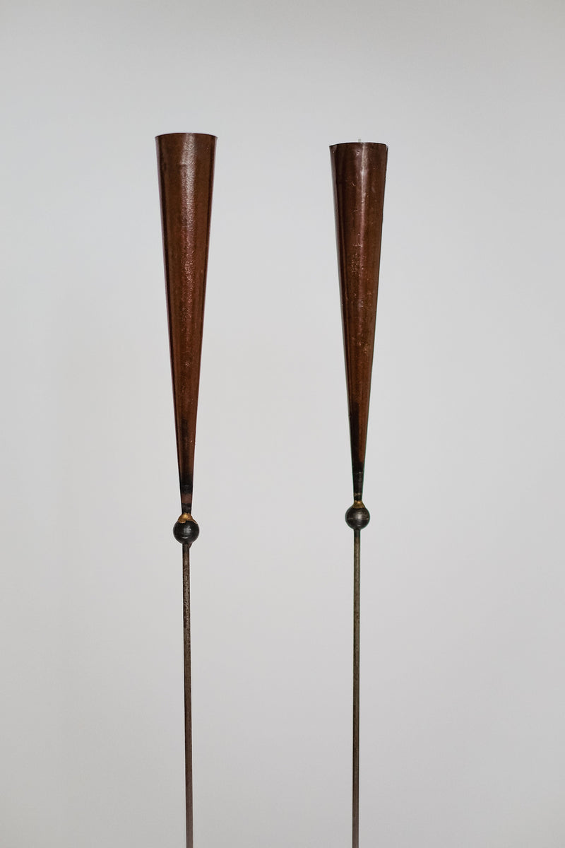 Two tall, slender, cone-shaped copper objects on thin rods with round base details are Pieter Spruyt Copper Candle Scones by septembre studios, shown against a plain white background.