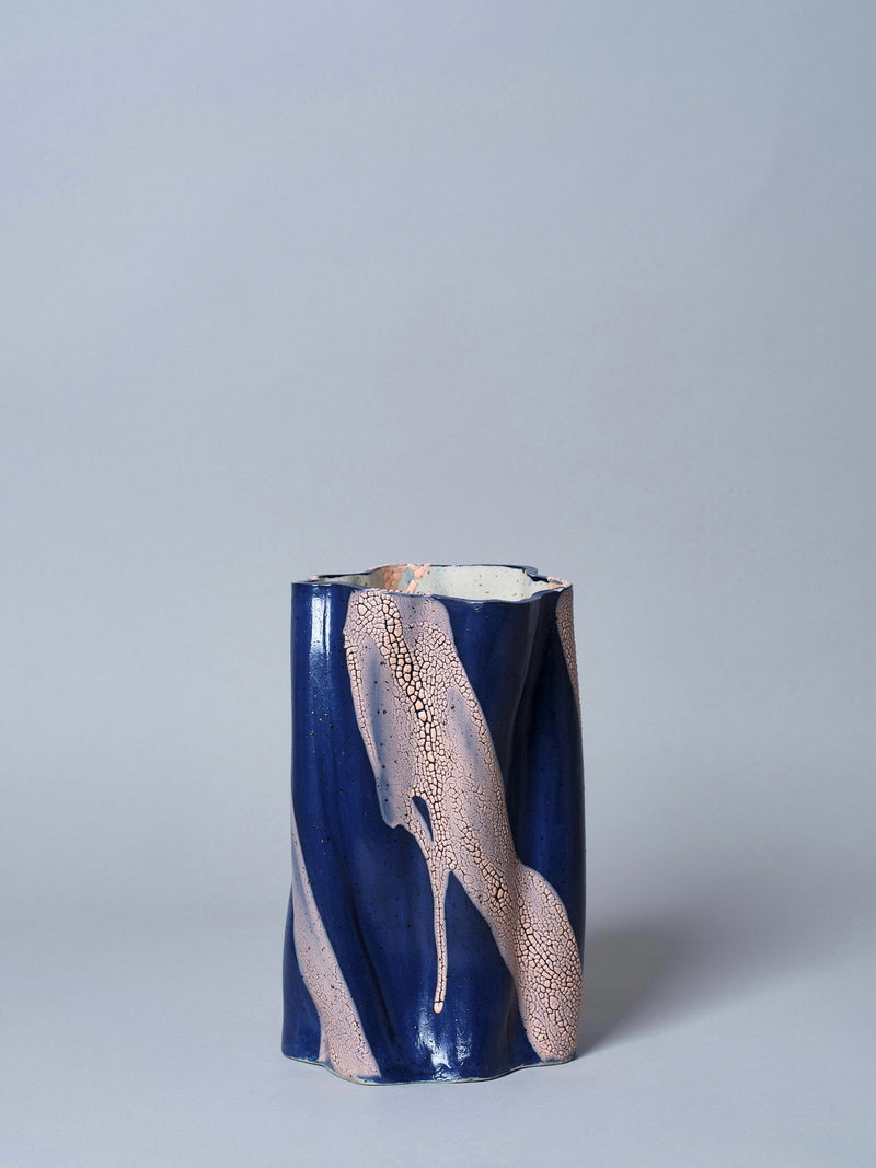 The Swirly Vase Blue and Peach by Astrid Öhman is a cylindrical handmade vase with a wavy surface, dark blue glaze, and light peach streaks, set against a plain light gray background—an artistic touch for any space.