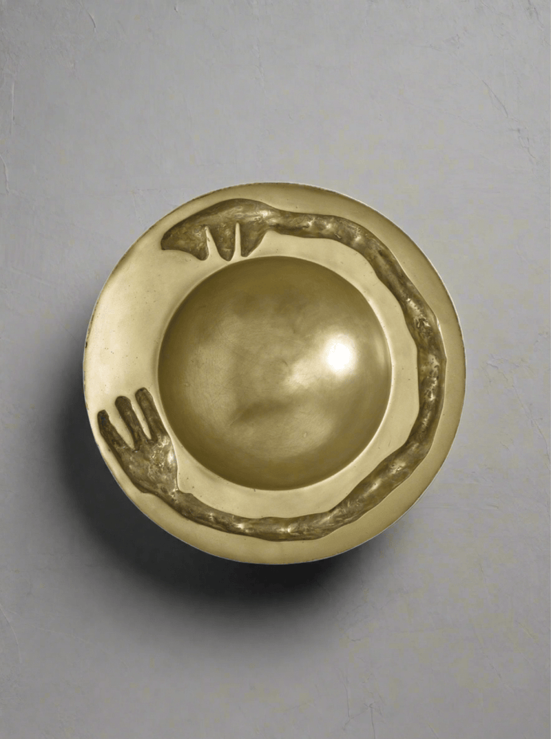 The Signature Dish Brass Sculpture by Lucy Page features a gold-toned, circular bowl with a sculpted snake forming a continuous loop along the raised rim—an eye-catching decorative piece on a smooth, light gray surface.