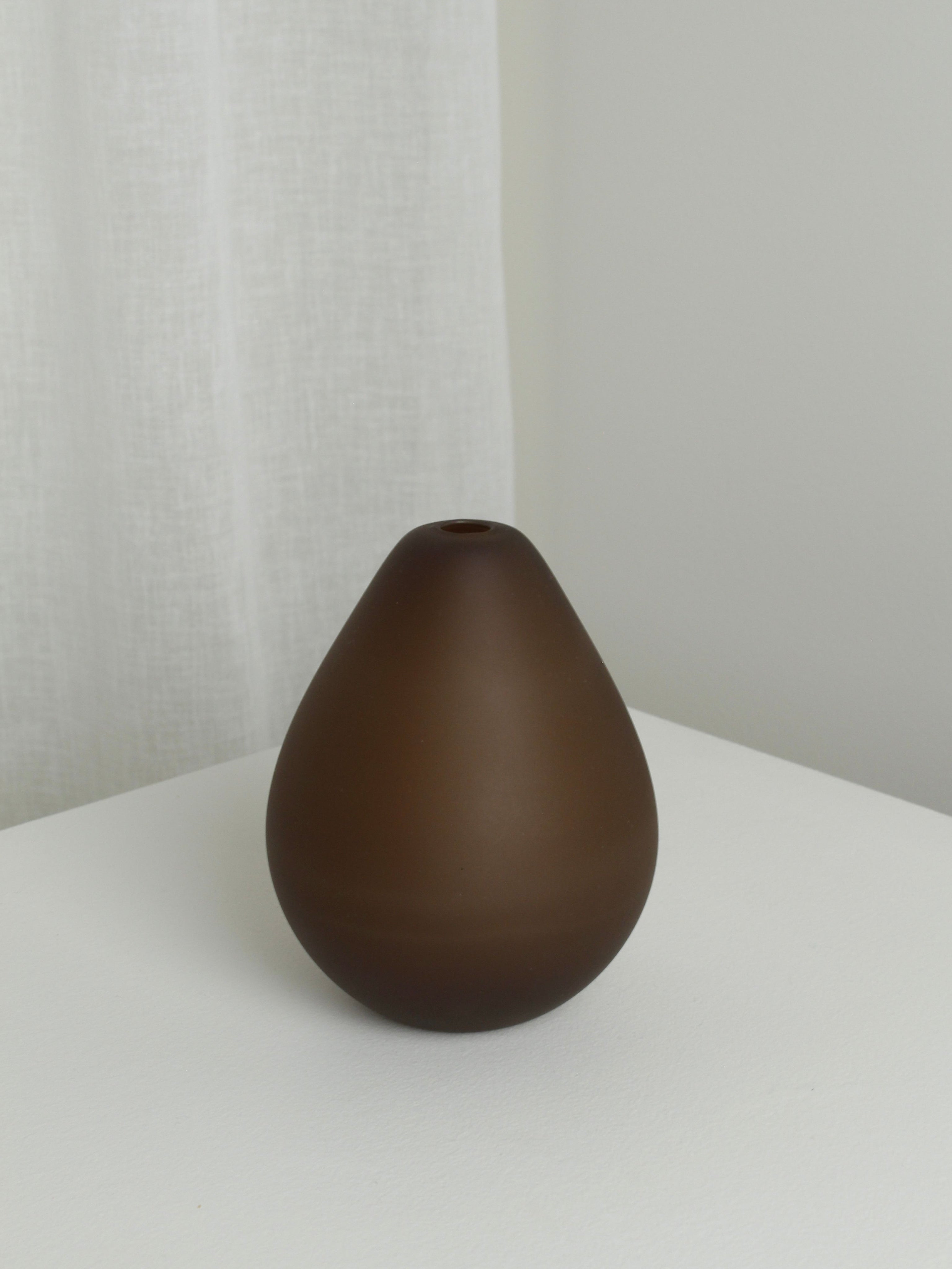 The Verre d'Onge Medium Vase in Chocolate features a matte teardrop design, displayed on a white surface with a light backdrop and white curtain, adding subtle warmth and elegance to any space.