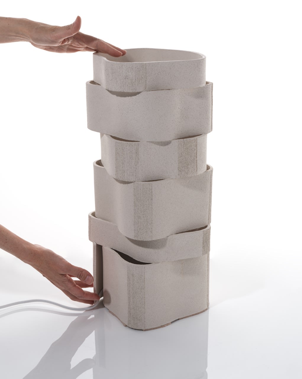 A person’s hands adjust the Katia Langeard 'Too 60' Ceramic Table Lamp, featuring four interlocking stacked sections and a power cord, against a white background.