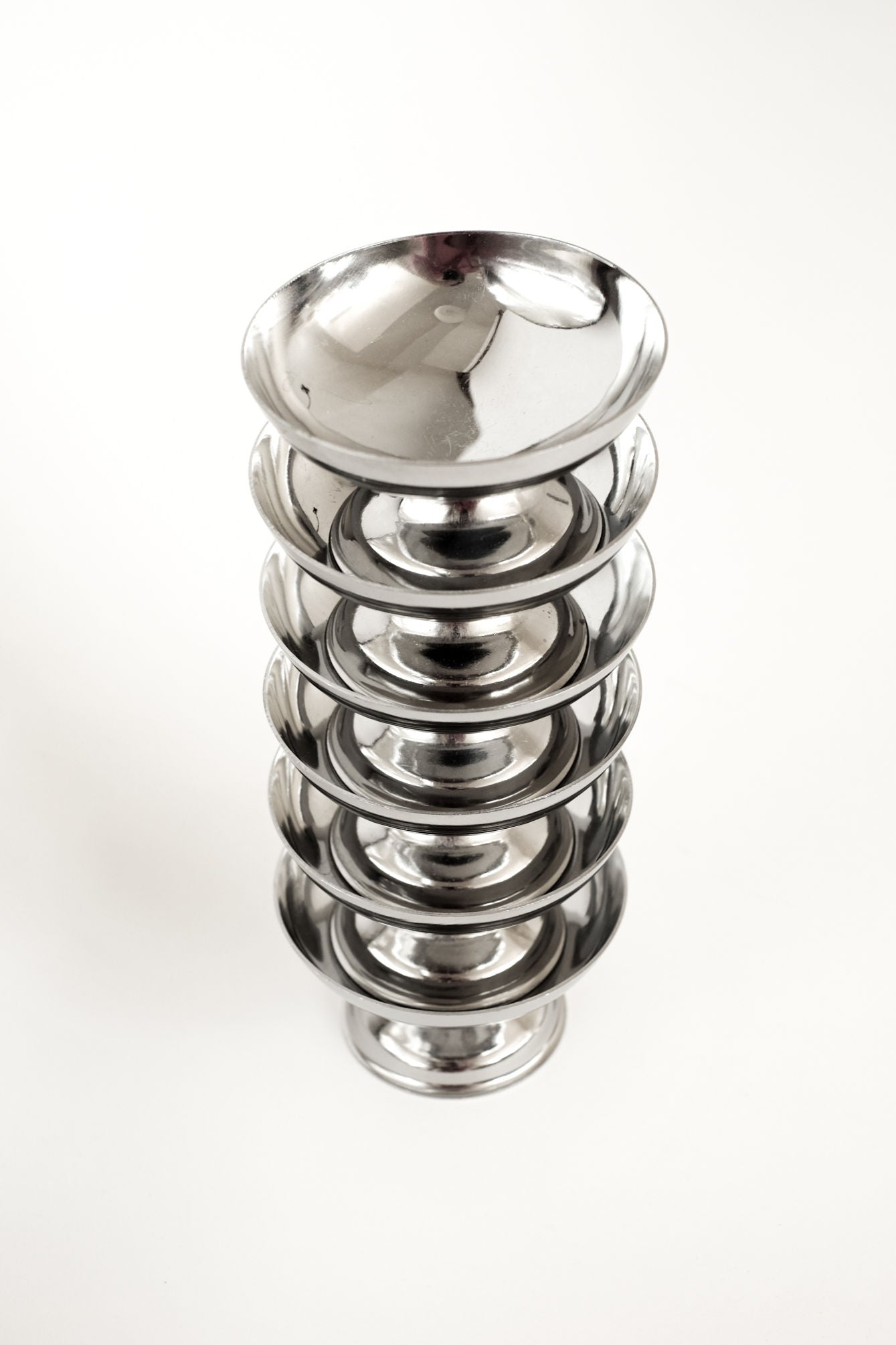 A set of six shiny, round stainless steel ice coupes by septembre studios are stacked vertically on a white background, reflecting light for a sleek, modern look.