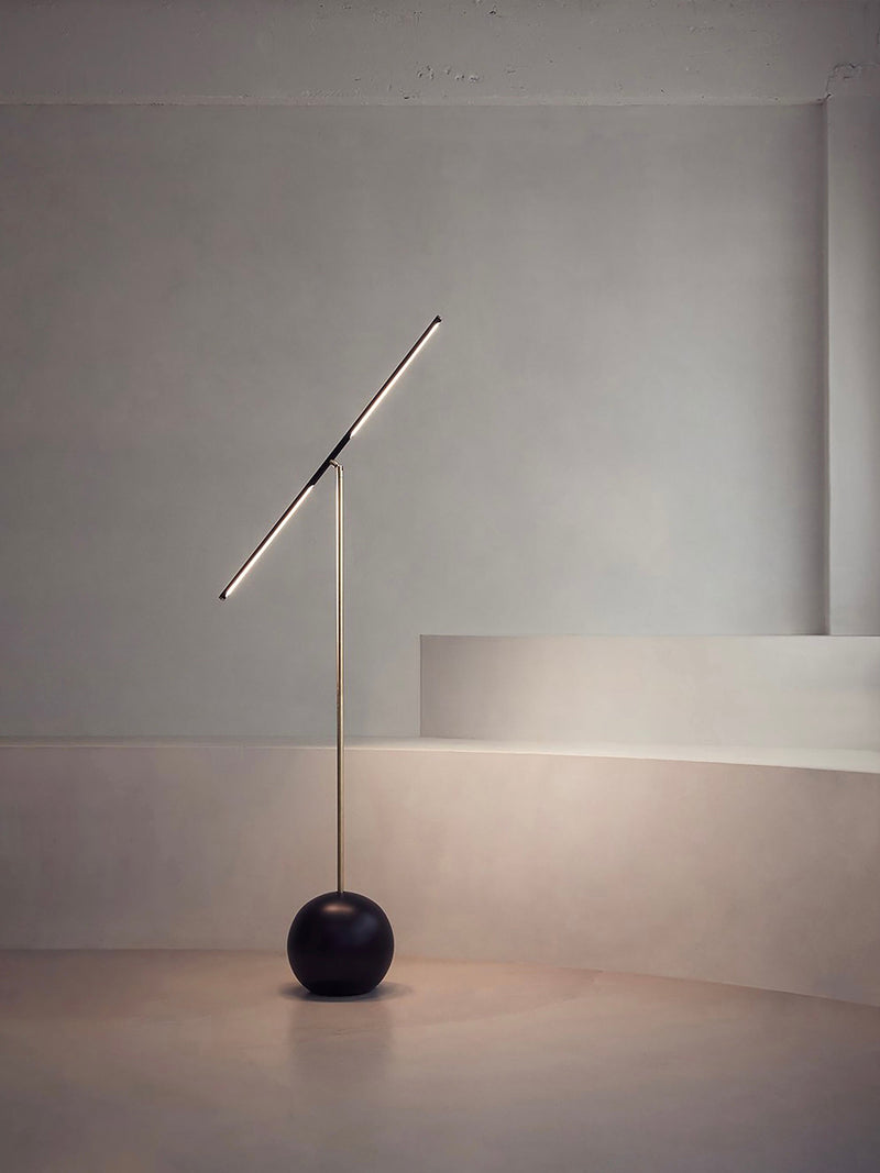 The 'T-Lamp' Floor Lamp by Heako Studio features a modern, sculptural design with a slim diagonal light bar on a slender pole and a round black base, perfect for neutral-toned rooms with curved steps and soft lighting.