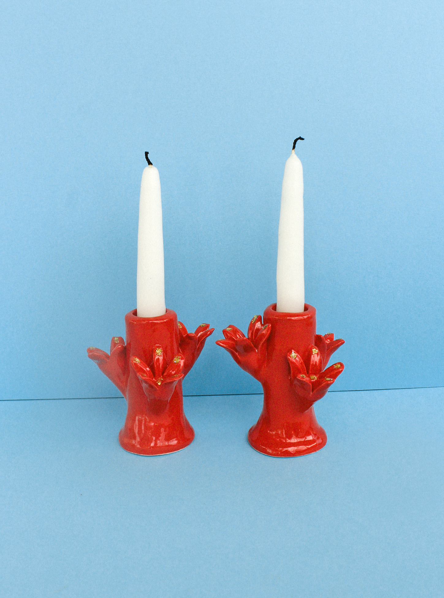 Sentence with replaced product and brand name: Two white candles, each standing in a Casa Veronica Flores Candleholder Set - Rojo shaped like a bird with outstretched wings, against a plain blue background.