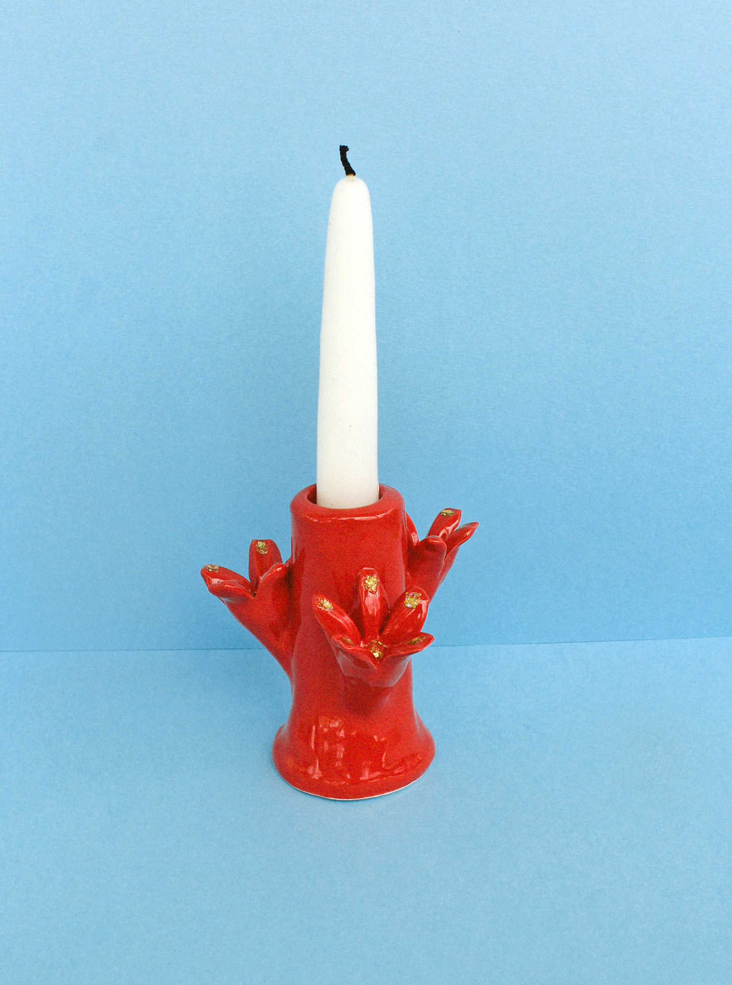 A white candle stands in a Flores Candleholder Set - Rojo by Casa Veronica, set with a hand design, against a plain blue background. The fingers of the holder are adorned with golden details.