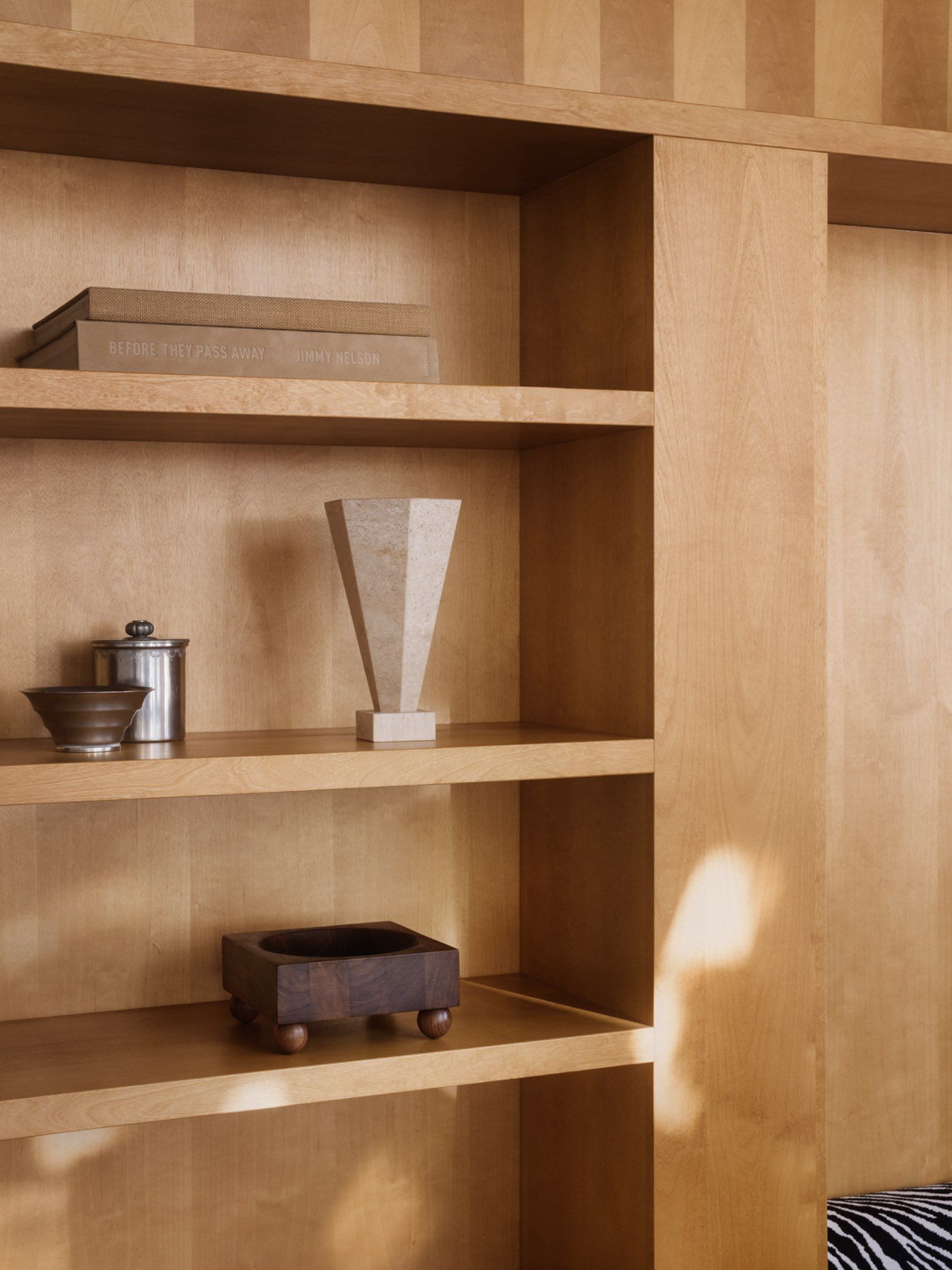 A wooden bookshelf with minimalist decor, featuring two stacked books, a metal container, Studio Vraco's Vase 01 Travertine from the TSDXSV Collection, and a dark wooden box with short legs. Soft sunlight patterns fall across the shelves.