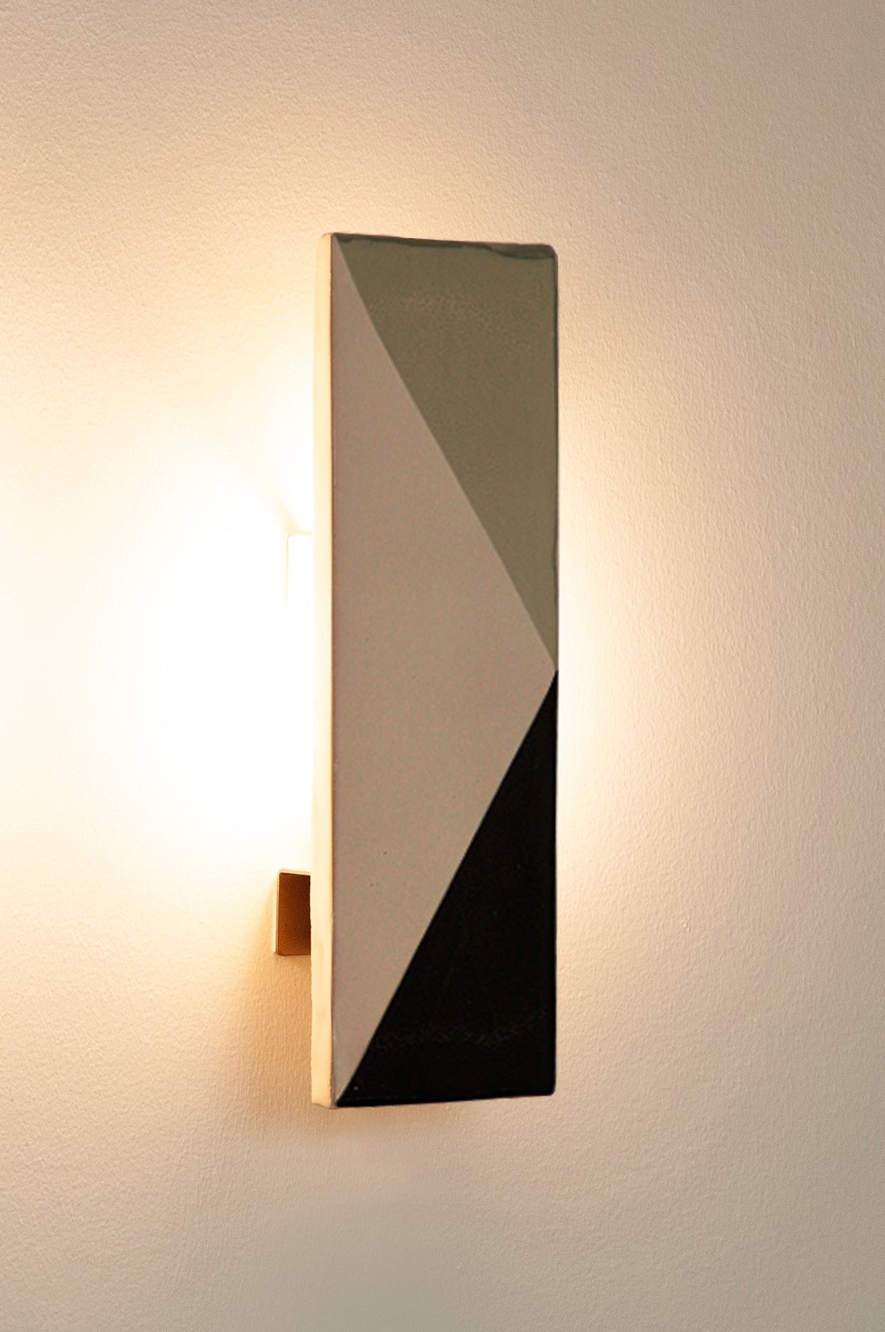 The Tile VN wall sconce by Violaine d'Harcourt features a modern rectangular geometric shade with black, gray, and white triangles. This artisan-made fixture adds elegance and emits soft light onto a plain, light-colored wall.