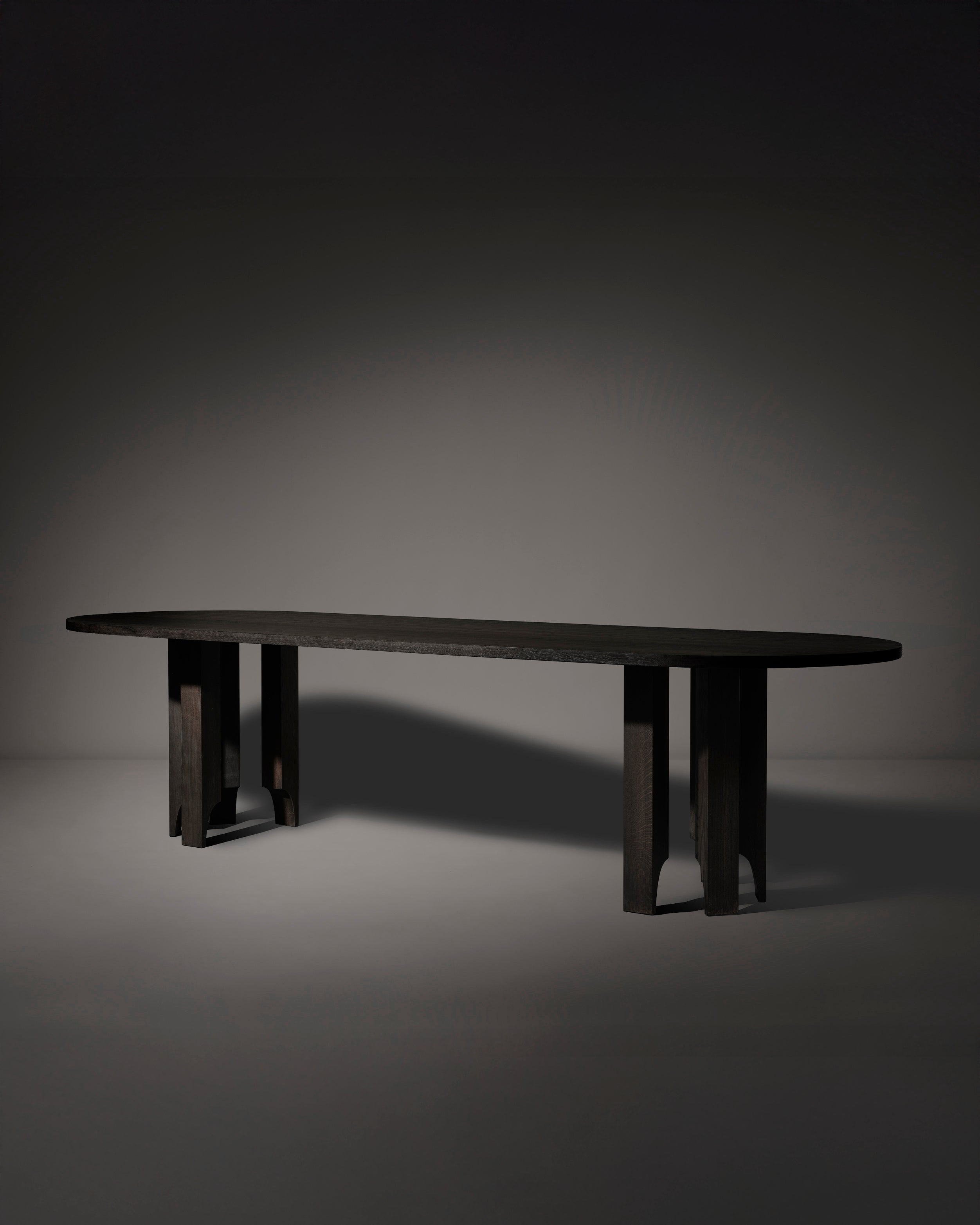 The Acer Capsule Table by Mokko is a long, oval piece made from solid European oak, featuring a smooth top and two wide geometric pedestal bases, shown against a dark neutral background.