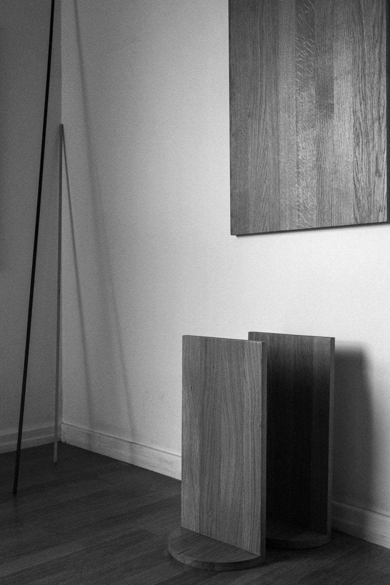 Minimalist black and white photo of a room corner featuring wooden flooring, a modern geometric wooden side table, two leaning sticks, a COSEINCORSO "Megaron" Shelf Black, and a wooden wall panel. The scene appears simple and uncluttered.