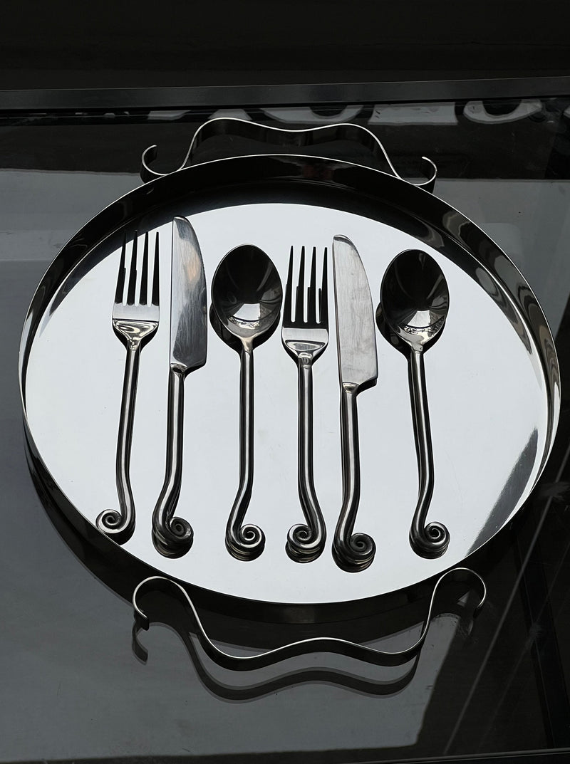 The Manushi Rare Vintage Swirl Cutlery Set—two forks, two knives, and a spoon with ornate spiral handles—is arranged neatly on a silver tray, showcasing their vintage charm on the reflective surface.