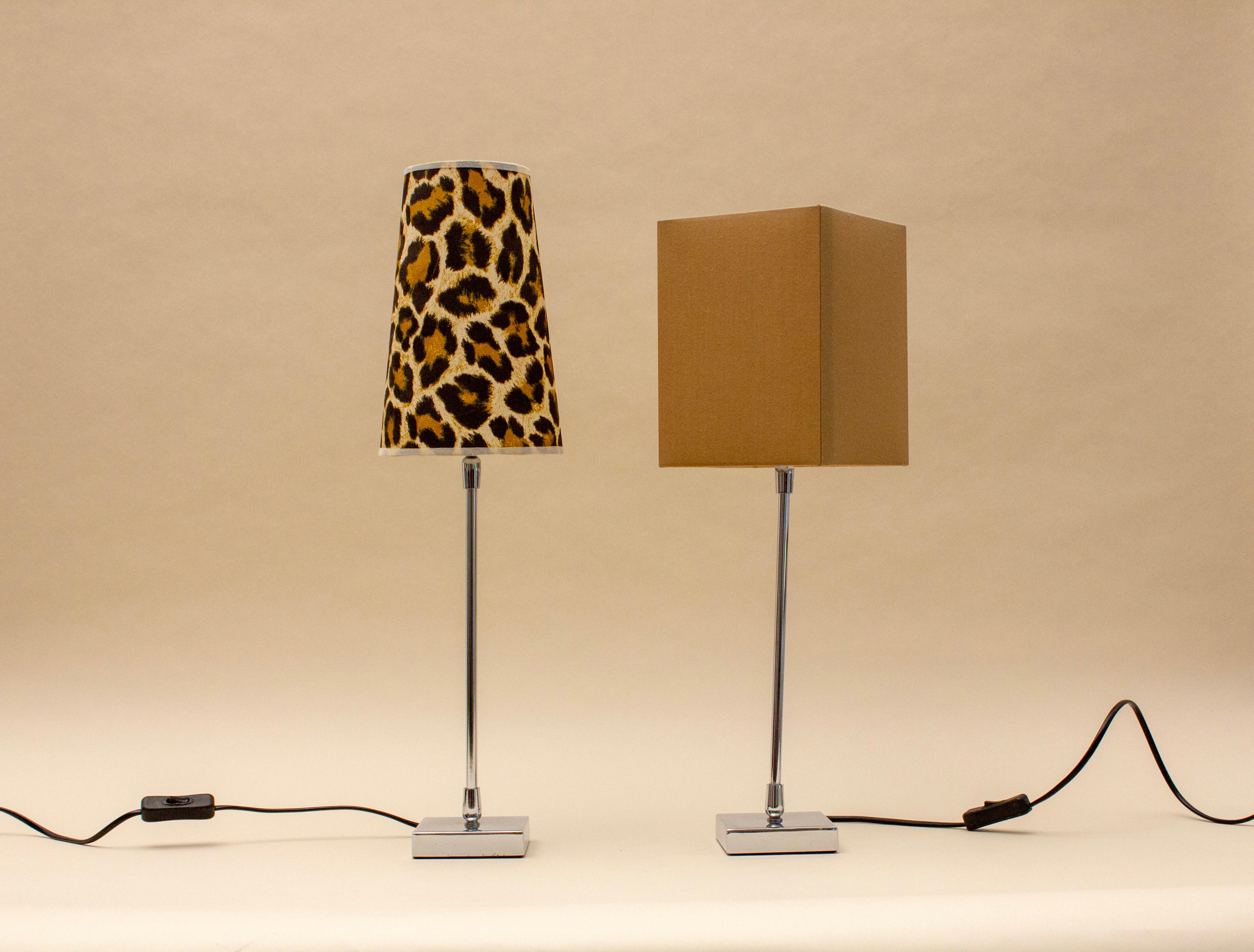 The SÒLER ZAFRA 2 TABLE LAMP set features two chrome-based lamps: one with a vintage conical leopard print shade and the other with a plain rectangular tan shade, both equipped with black cords and switches.