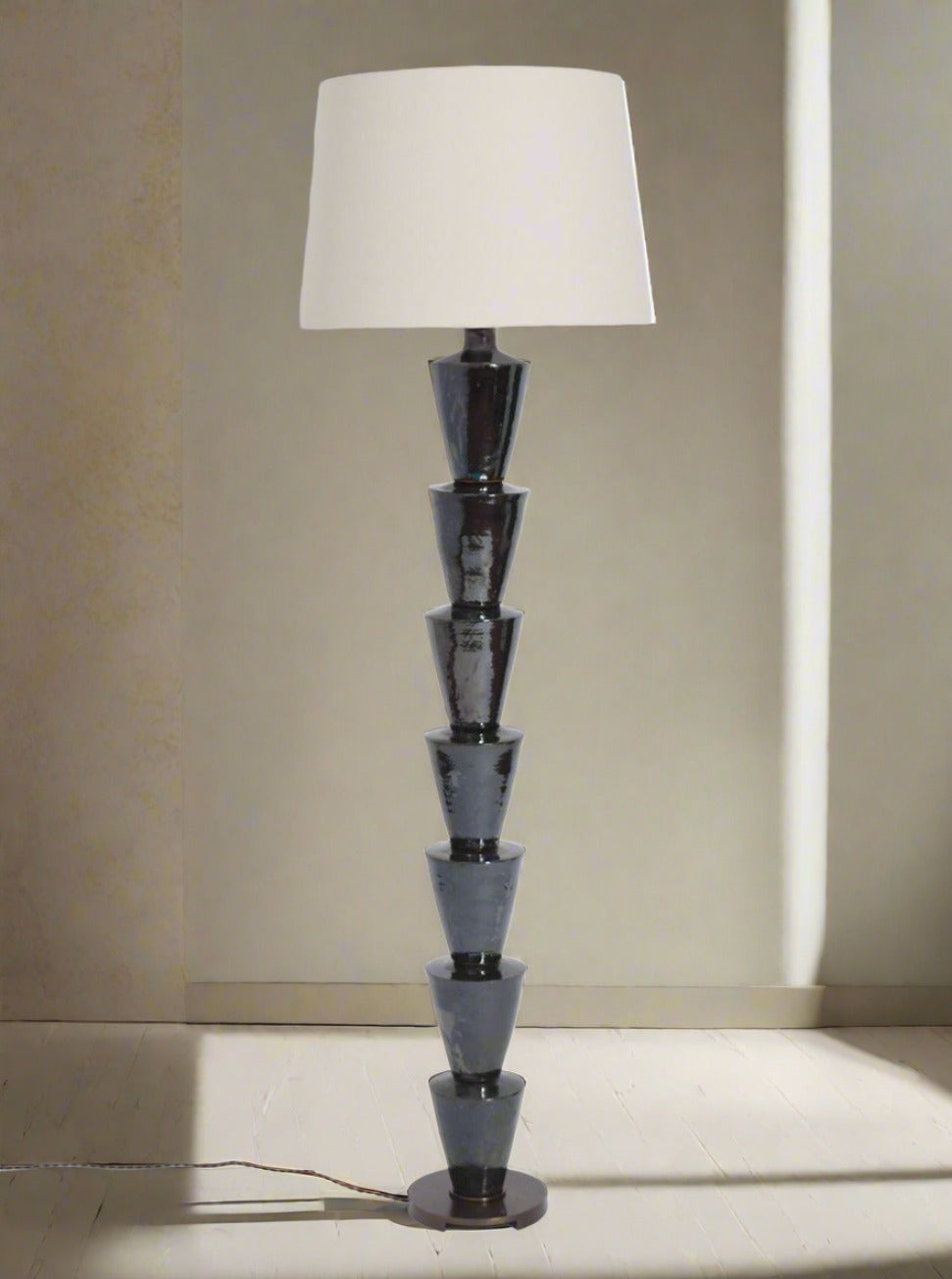 The "Nizwa" Floor Lamp by Barracuda Interiors features a modern, glazed stacked geometric black base and a large white shade, standing on a light wooden floor and delivering customizable lighting enhanced by natural side light.