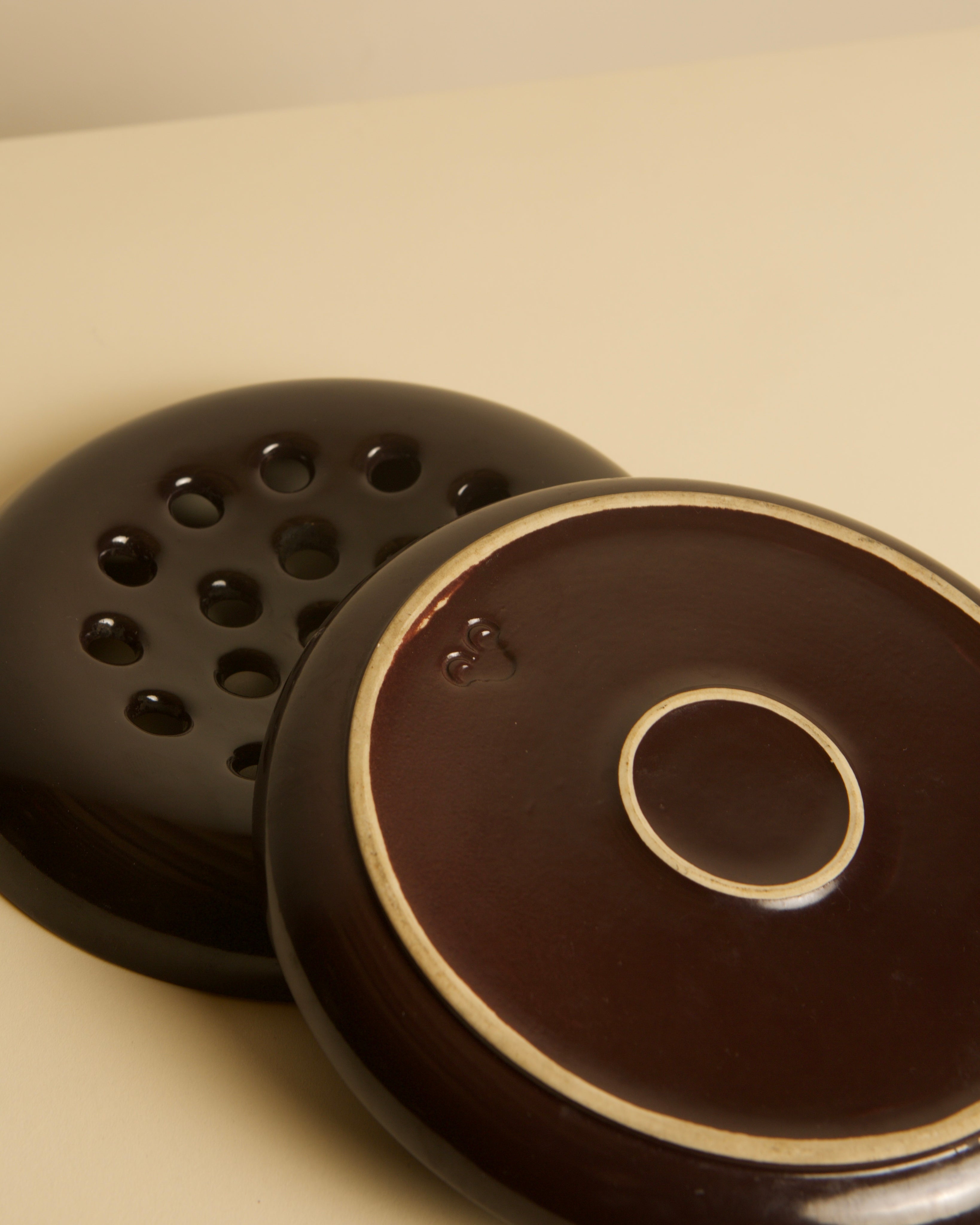 Two Italian brown ceramic ashtrays by Angelo Mangiarotti for Brambilla (1960s) from Treaptyque are shown: one with multiple holes, the other solid with a raised circular center, both resting on a light surface.