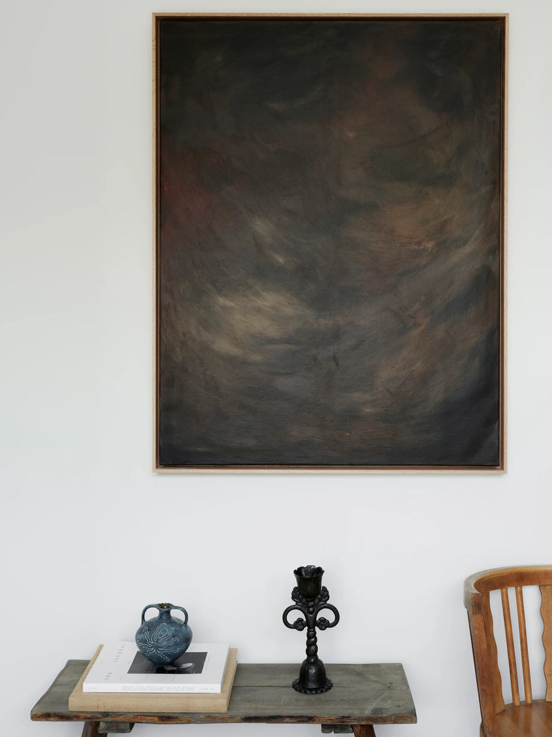 A dark, abstract painting titled "Shadowed Abyss - Mixed media on canvas" by Mattias Storm hangs on a white wall above a small table. The table holds a stack of books, a blue ceramic vase, and a decorative black candle holder. Part of a wooden chair is visible to the right.