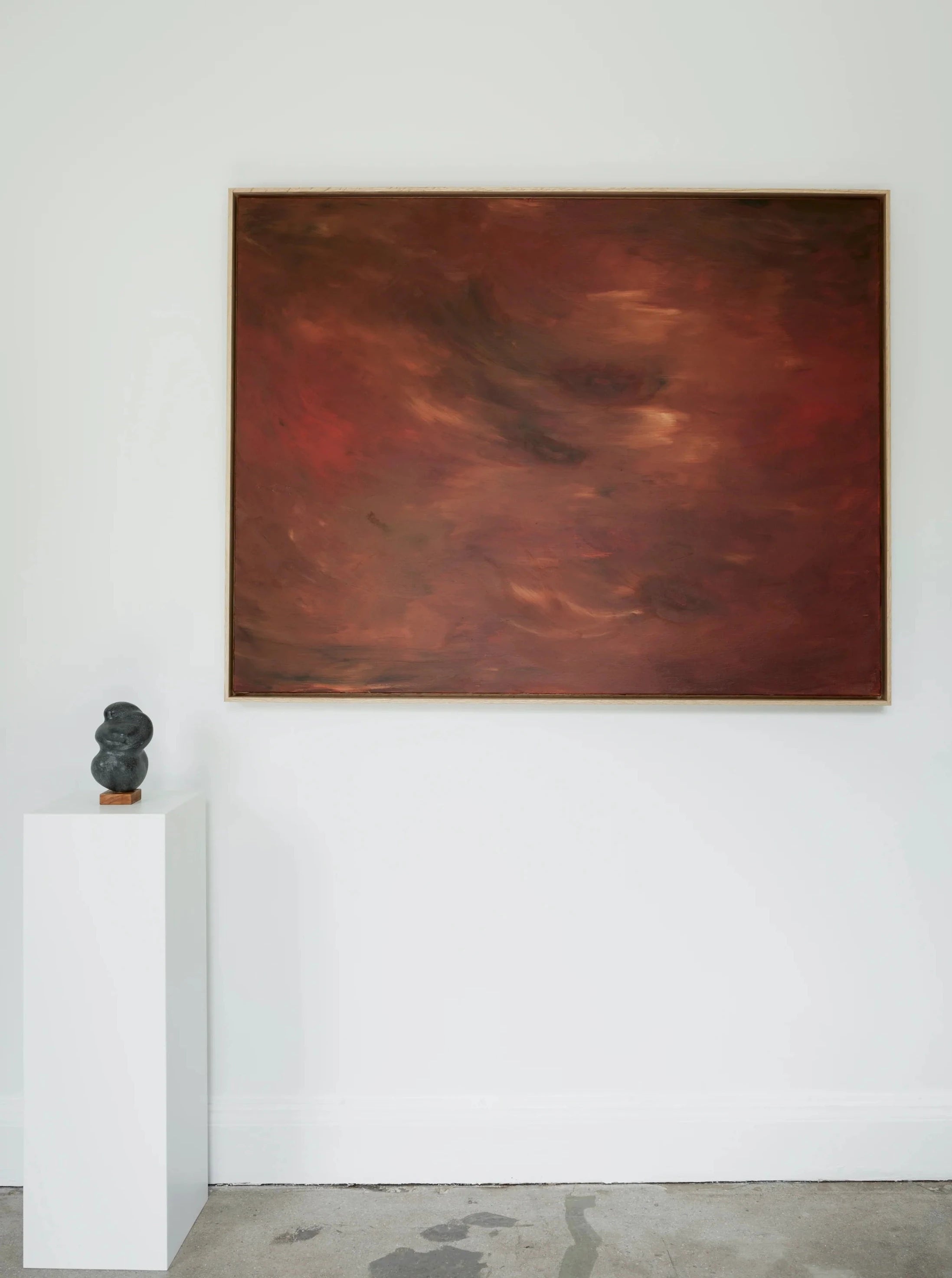 A minimalistic gallery display features "Crimson Horizons" by Mattias Storm, a large abstract painting with red and brown hues from the Terra Incognita series. The artwork is mounted on a white wall in an oak frame. Below and to the left, a small dark-colored sculpture rests on a white pedestal, enhancing the clean, modern aesthetic of the room.