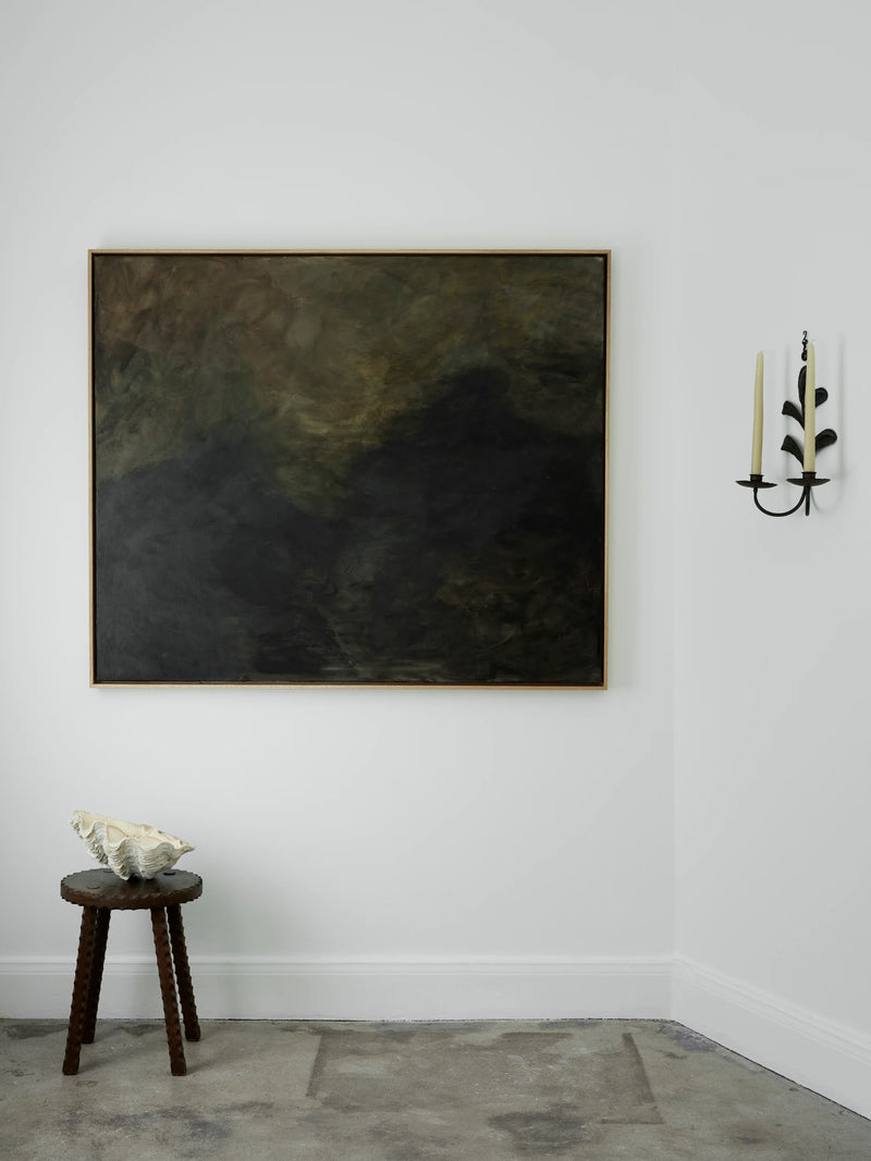A minimalist room features a large abstract painting titled "Veiled Dusk - mixed media on canvas" by Mattias Storm with dark, earthy tones on a white wall. The artwork is encased in an elegant oak frame. Below it, a small wooden stool with a seashell rests quietly. To the right, an unlit candle sits in a leafy design holder mounted on the wall.