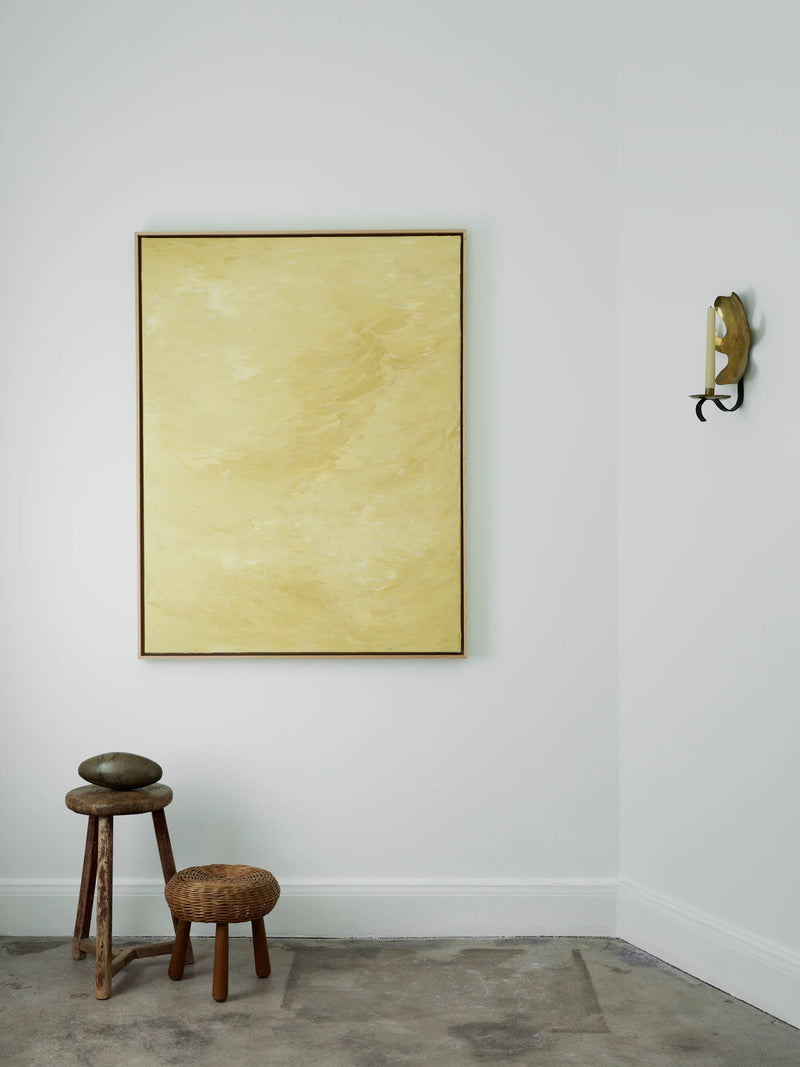 A minimalist room features a large framed yellow painting on a white wall, Solar Mirage - Mixed media on canvas by Mattias Storm. Below, two wooden stools of different heights sit against the wall. A round stone is placed on the taller stool. A single wall sconce with a candle extends from the wall to the right.