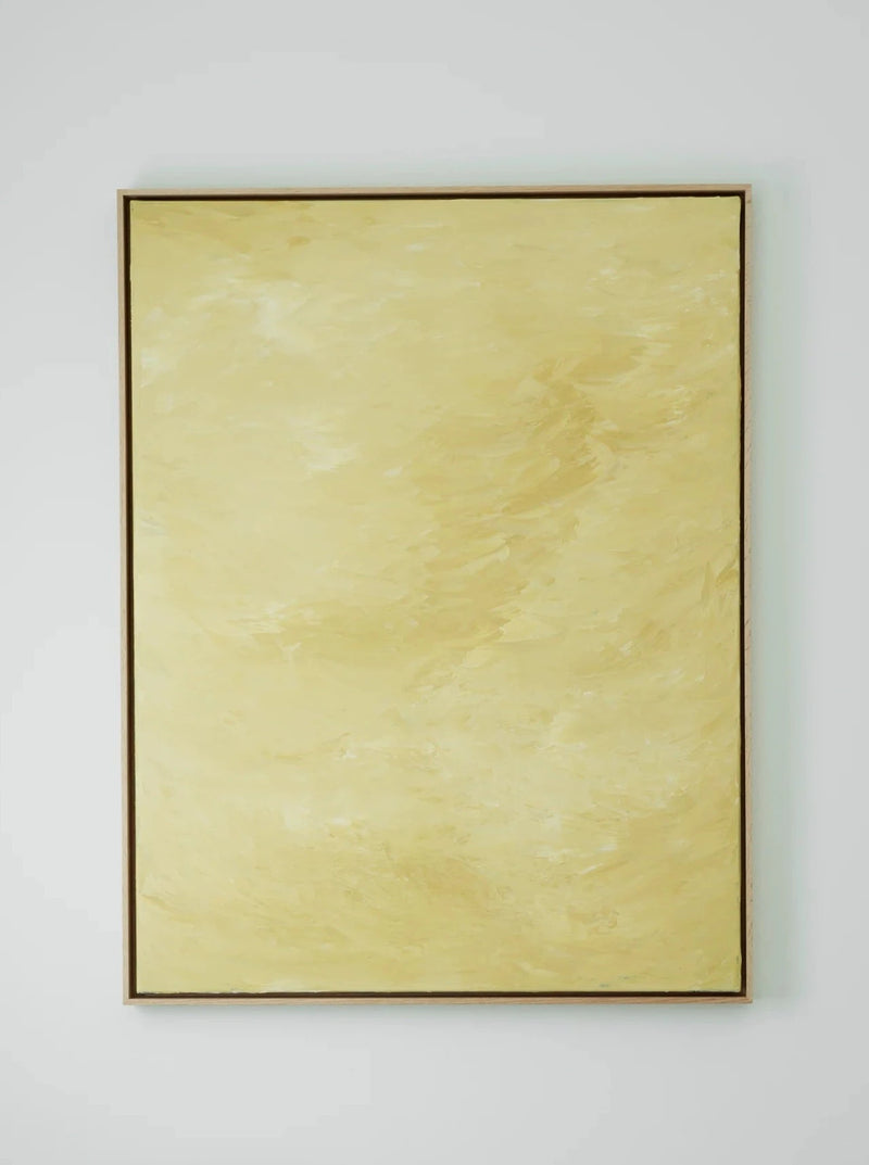 A minimalistic room featuring a large, framed yellow abstract painting titled "Solar Mirage - Mixed media on canvas" by Mattias Storm on a white wall. Below the artwork are two small, rustic wooden stools. One stool holds two balanced stones. A candle sconce with a candle adorns the wall to the right.