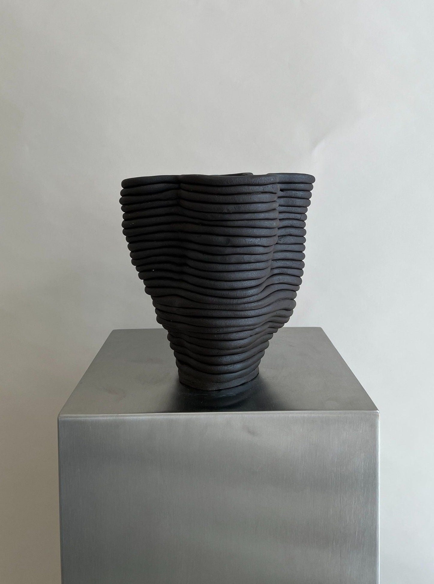 The Wobbly Vase Black by 2222Studio is a handcrafted ceramic piece with dark, wavy, stacked coils and an irregular texture. Presented on a metallic pedestal, it serves as an elegant blossom holder with a wide opening against a plain background.