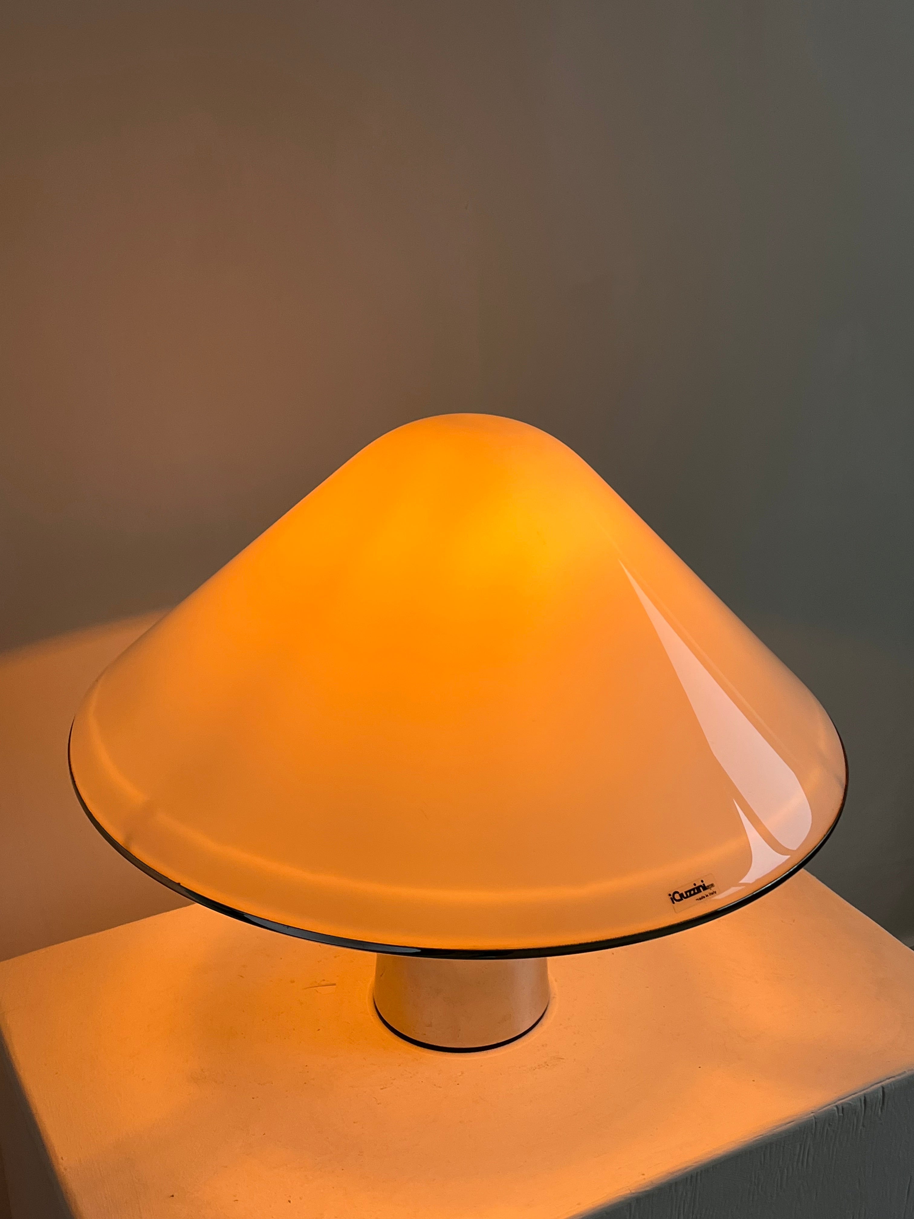 The Harvey Guzzini Elpis Table Lamp by septembre studios features a modern mushroom shape with a glossy beige shade, casting a warm orange glow on a white surface against a neutral wall.