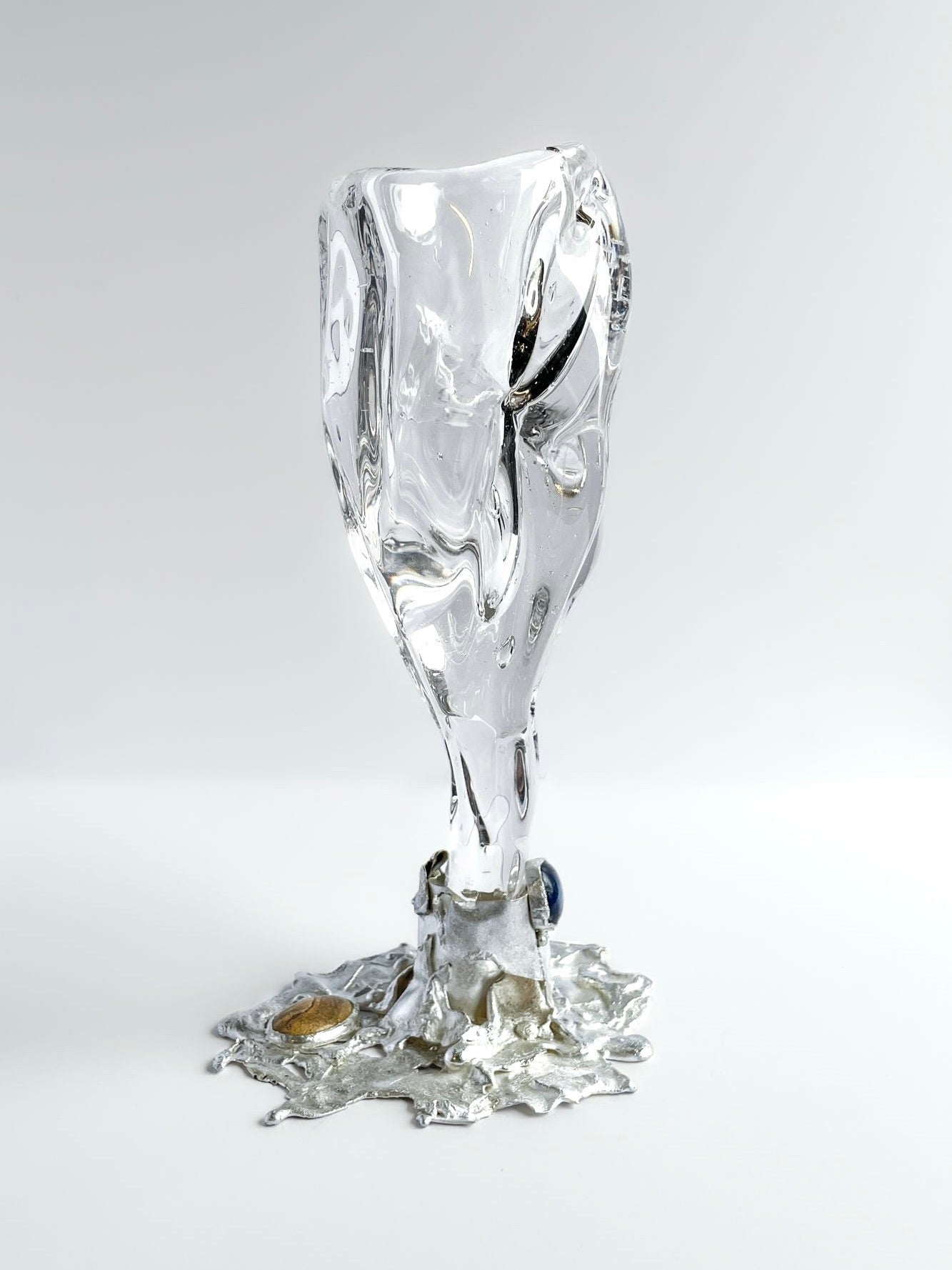 The Jewelled Goblet no. 5 by Szkło Studio x Lindsey Fontijn features a distorted, hand-blown glass form with a silver base spreading unevenly and embedded stones, shown on a flat surface against a white background.