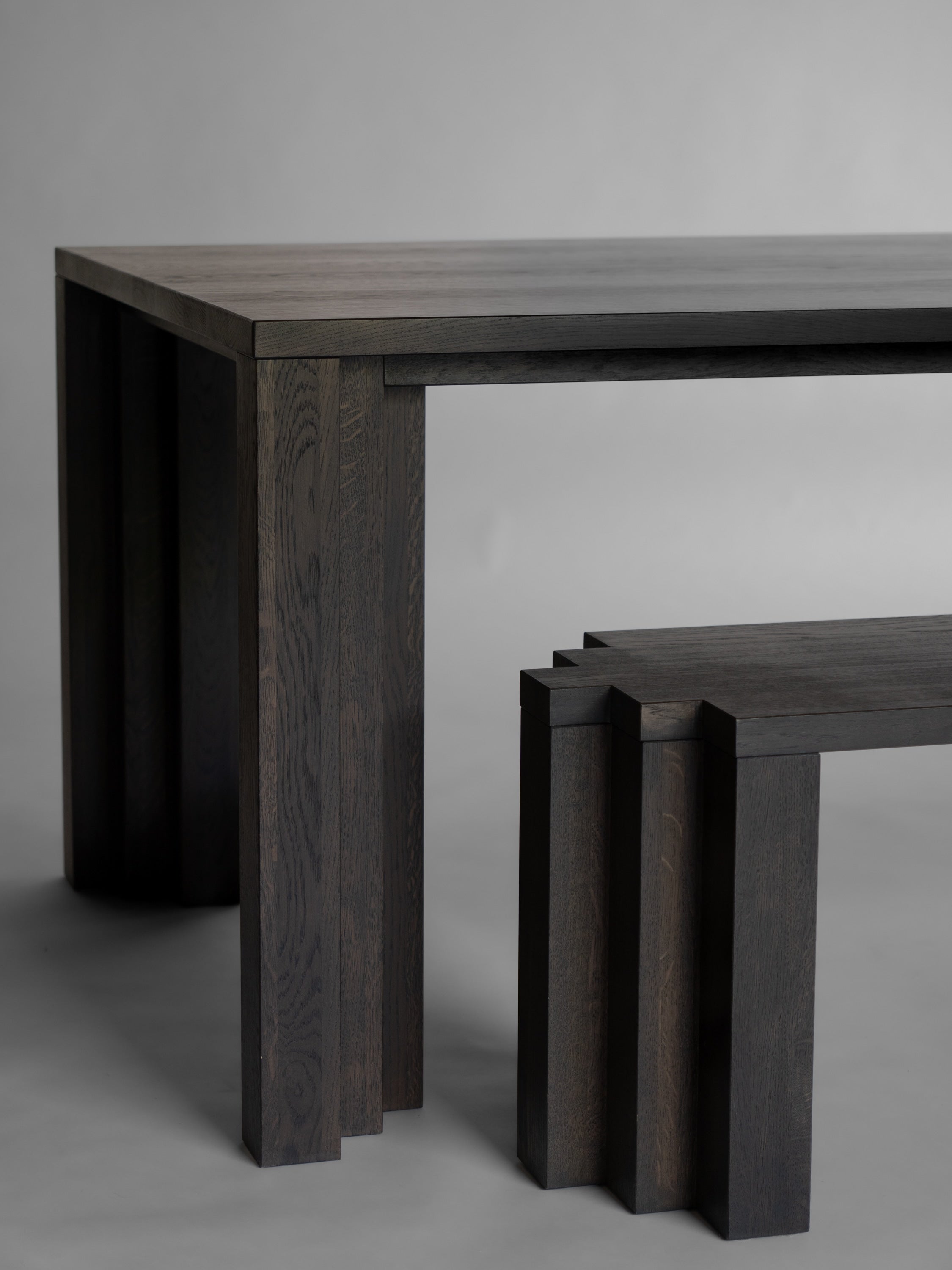 The Mokko Cadence Bench features thick geometric legs and solid European oak construction. This modern, minimalist piece pairs perfectly with a dark wood table, offering timeless, architectural appeal against a plain gray backdrop.