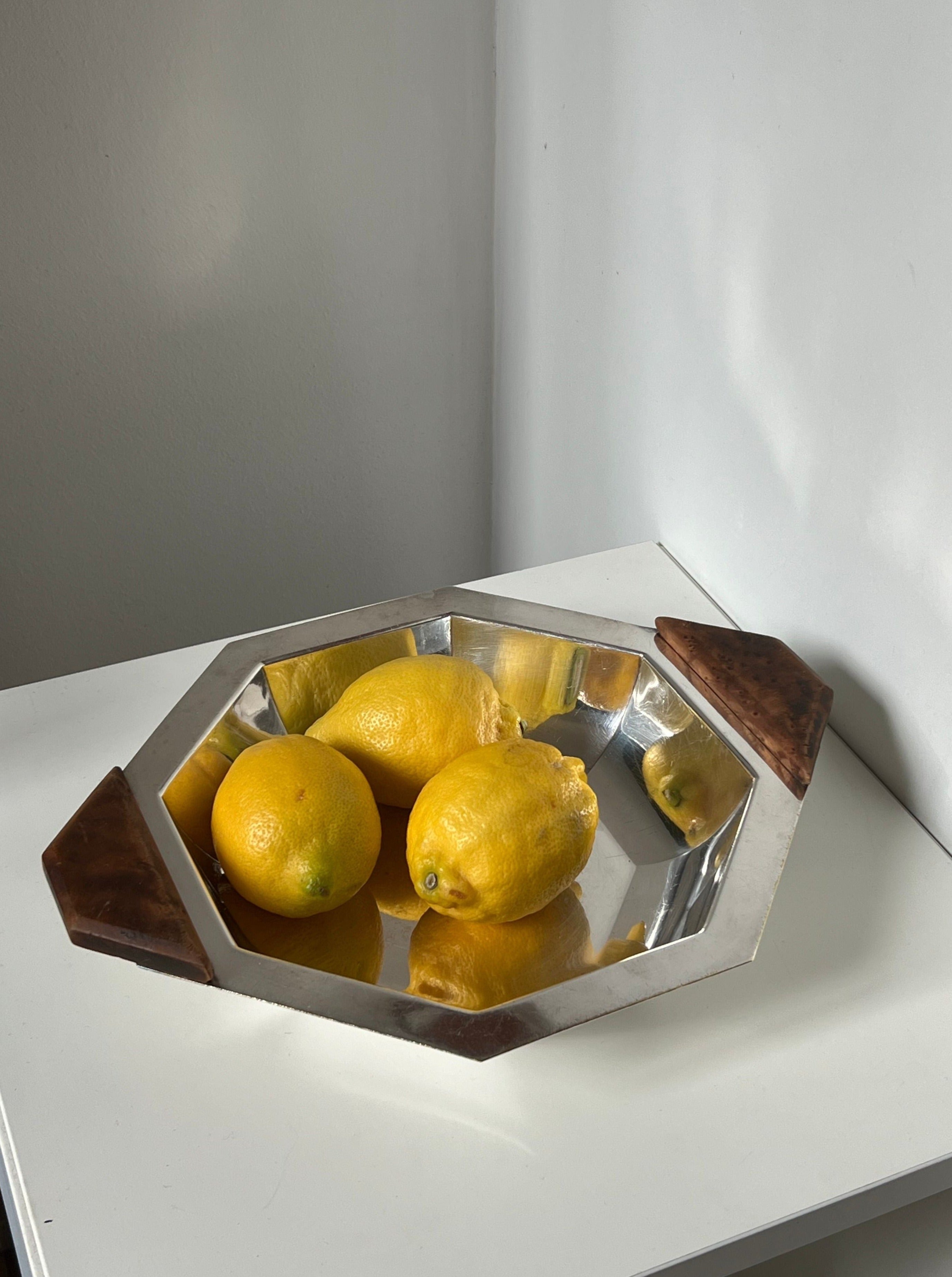 The Manushi Art Deco Decorative Dish, a silver octagonal piece with wooden handles, holds four yellow lemons on a white surface in the corner of a light-colored room.