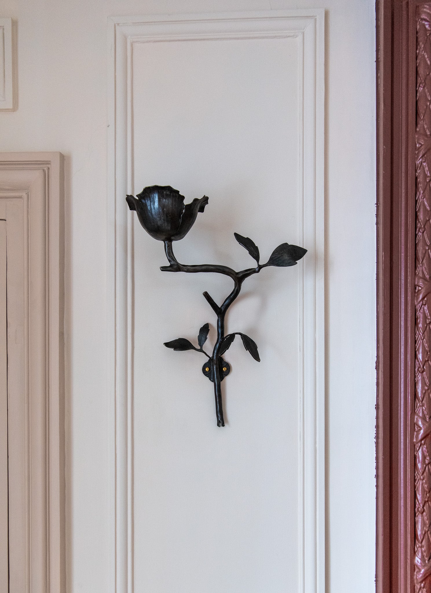 The Clematis Wall Light by Jess Wheeler Studio, a black handcrafted forged iron fixture shaped like a flower with leaves, is mounted on a white paneled wall next to a beige door and maroon textured surface.